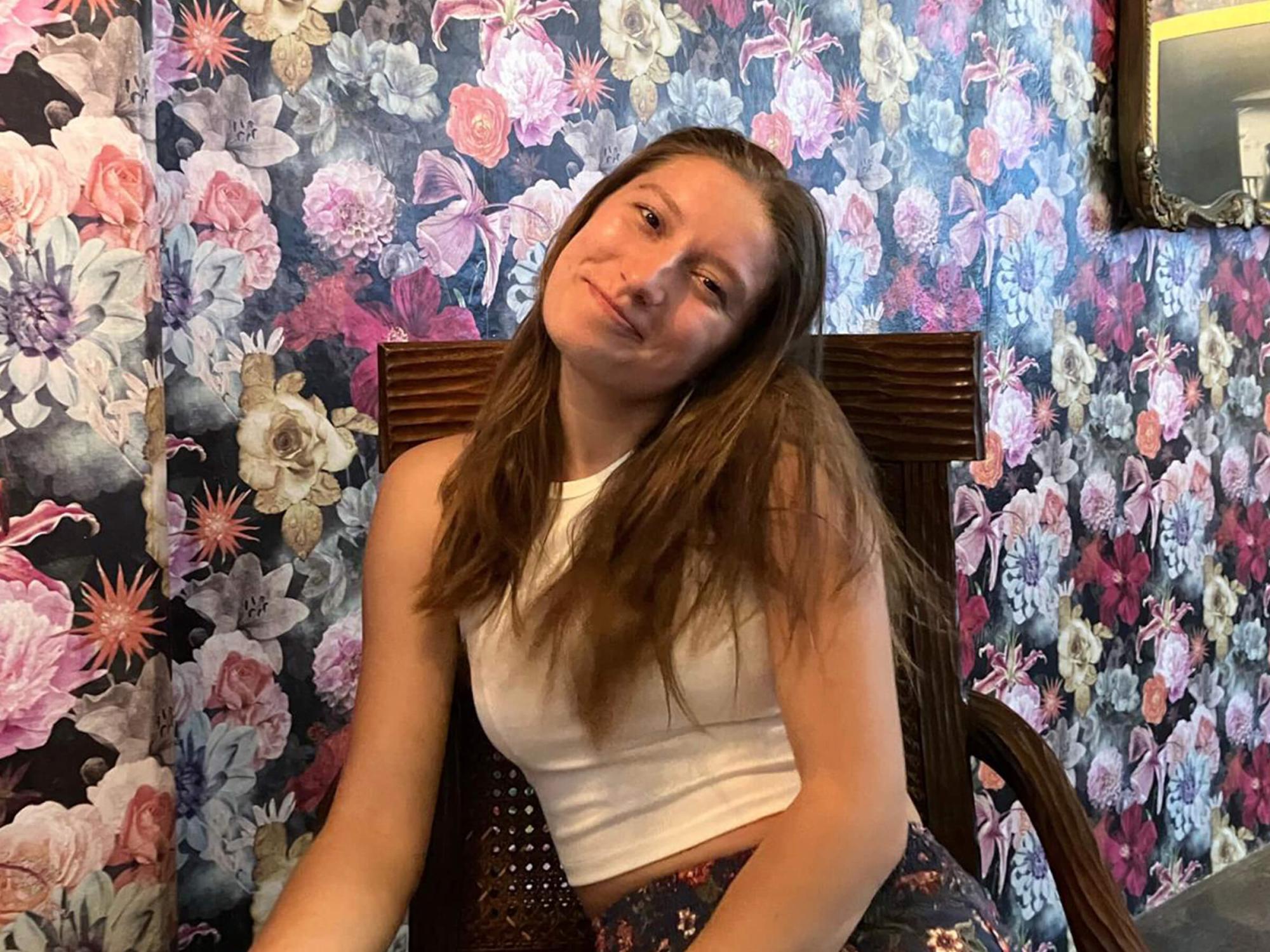 Anna Raquel Duiker sits in a chair in front of a wall covered in floral wallpaper.