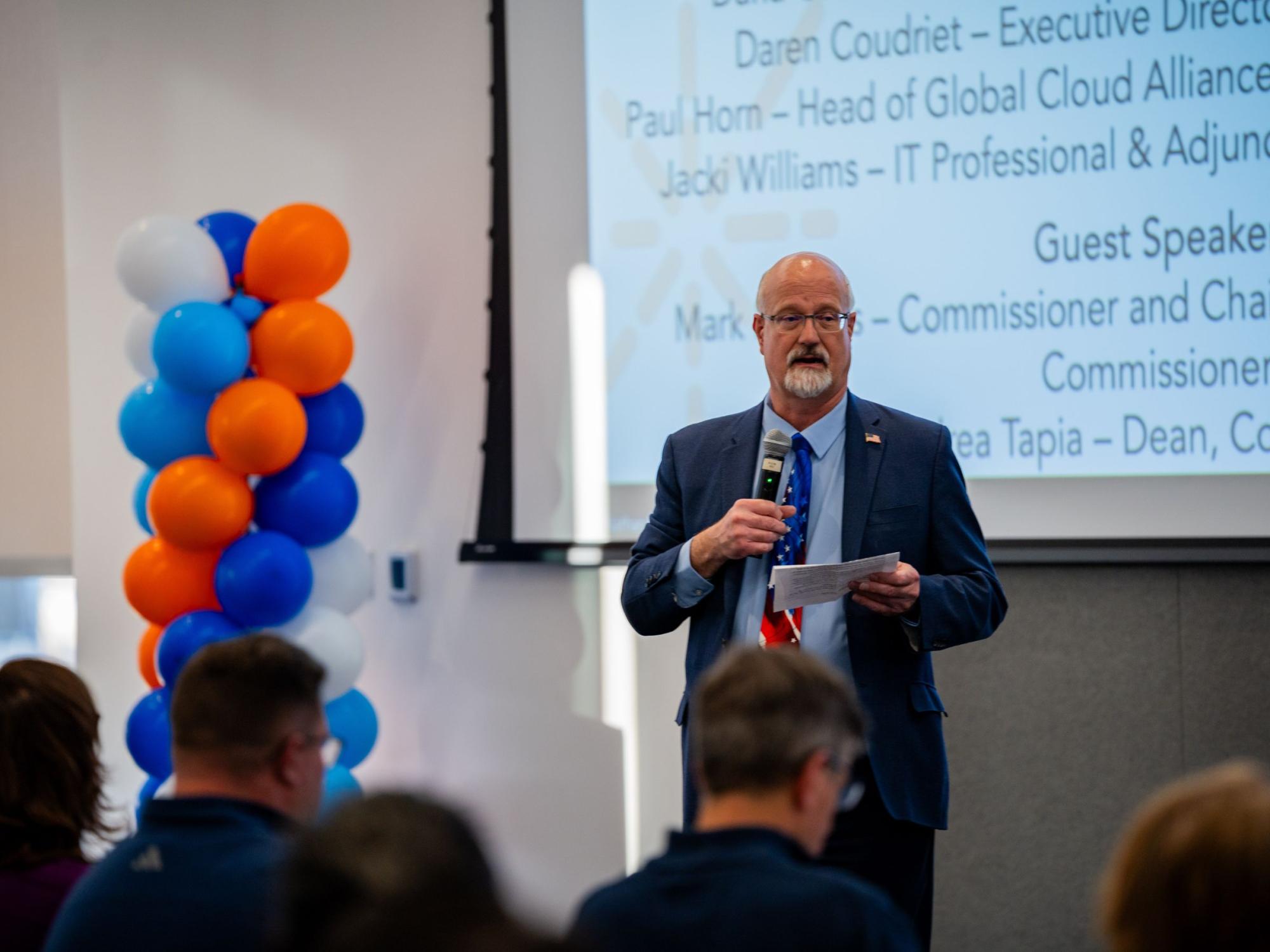 bald person with glasses and white facial hair speaks into microphone next to balloon display