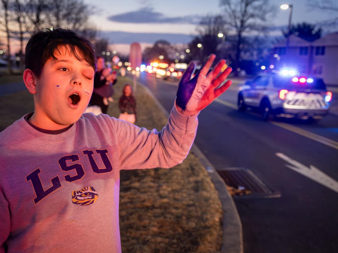 A young boy wearing an LSU sweatshirt waves to vehicles with flashing lights as they drive by. His hand is painted red, white and blue.
