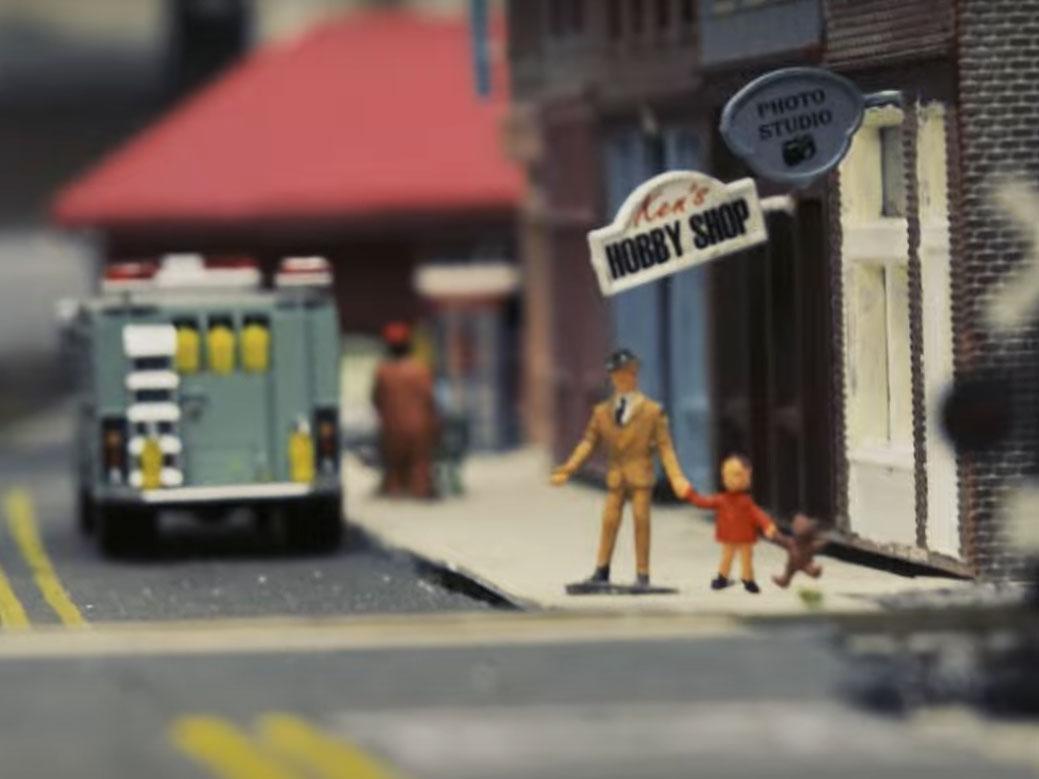 Two tiny figures are posed at an intersection in a model railway's city.