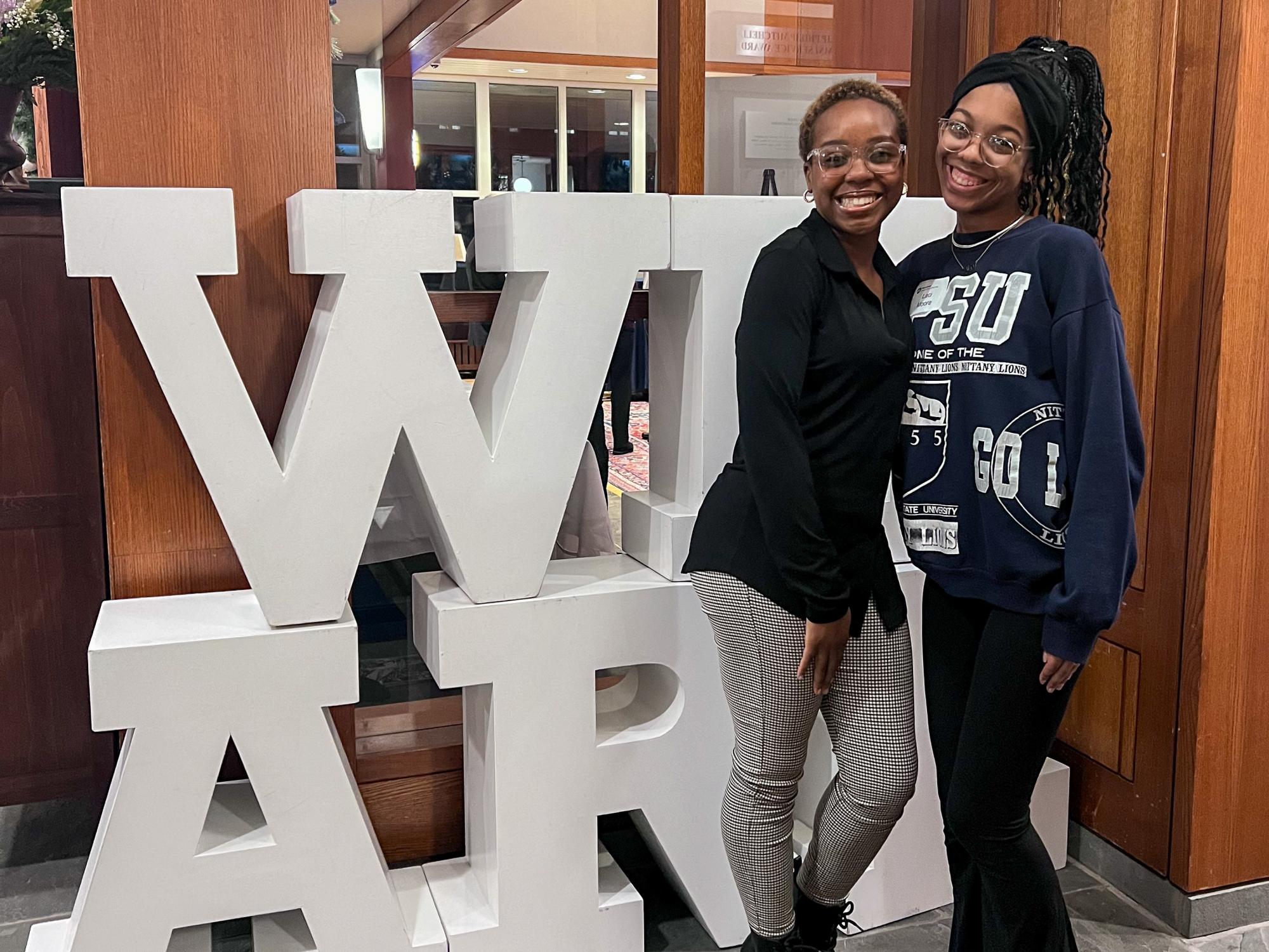 Laci Campbell (right) with her Chaiken Scholars mentor Nia Smith (left) at the 2023 Chaiken Scholars Reception in front of large white letters spelling WE ARE in the Hintz Family Alumni Center.