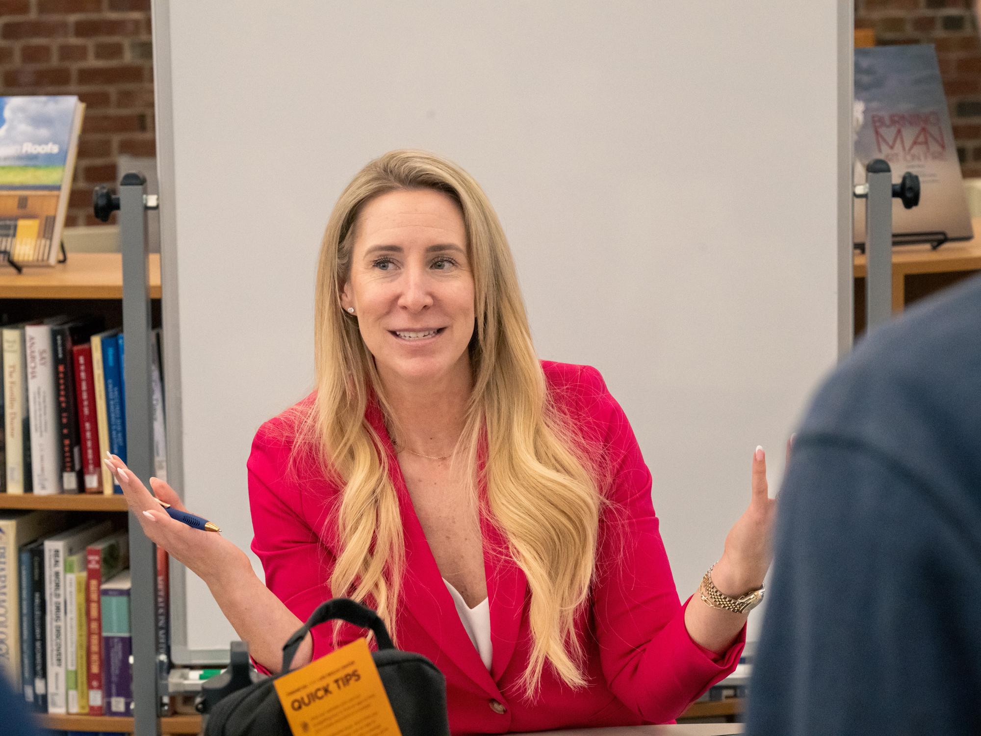 Erin Weber, dressed in a bright pink blazer, gestures speaking to students at a table in the library.