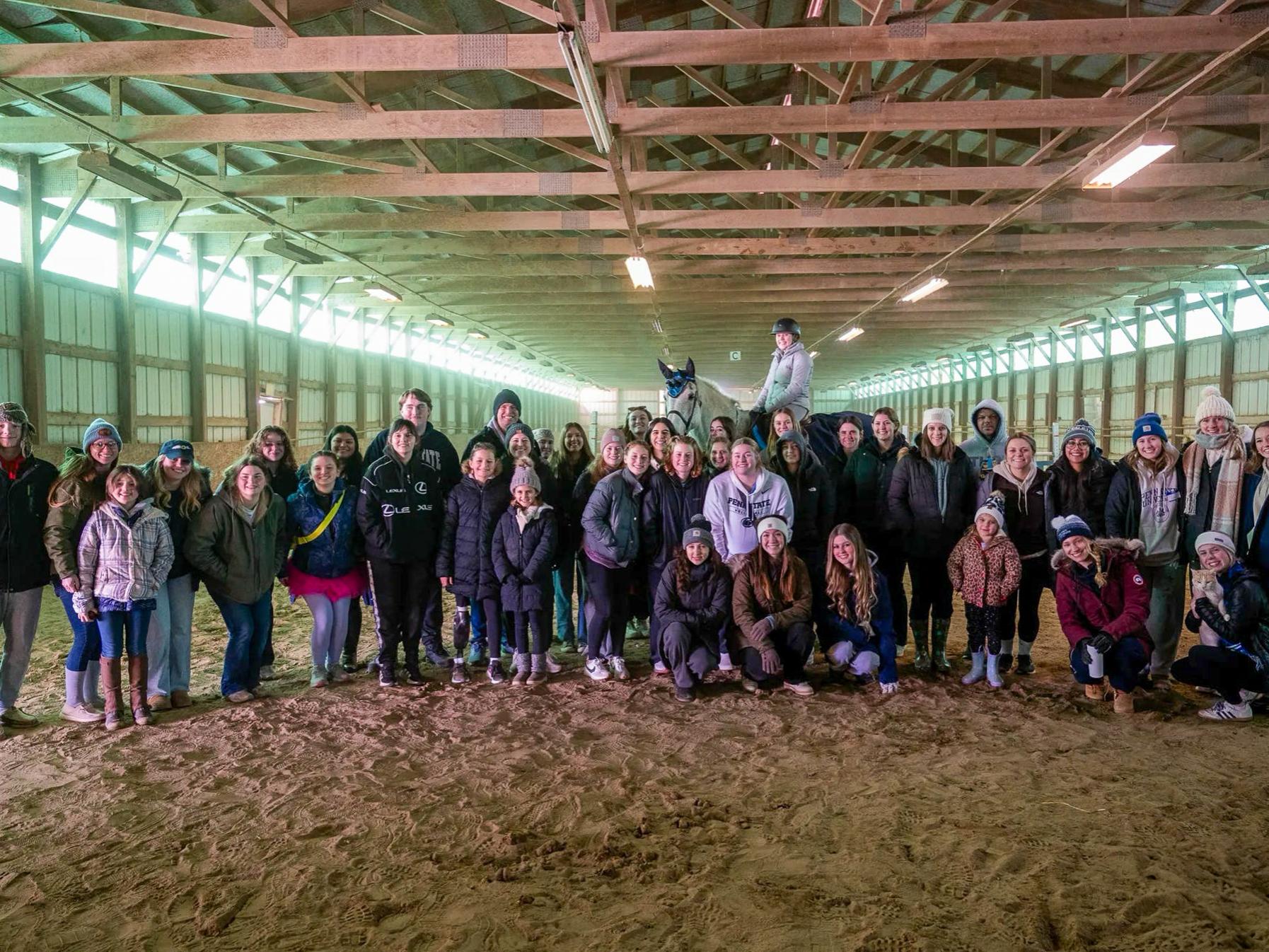 A group of people gathered in a barn with a horse in the back.