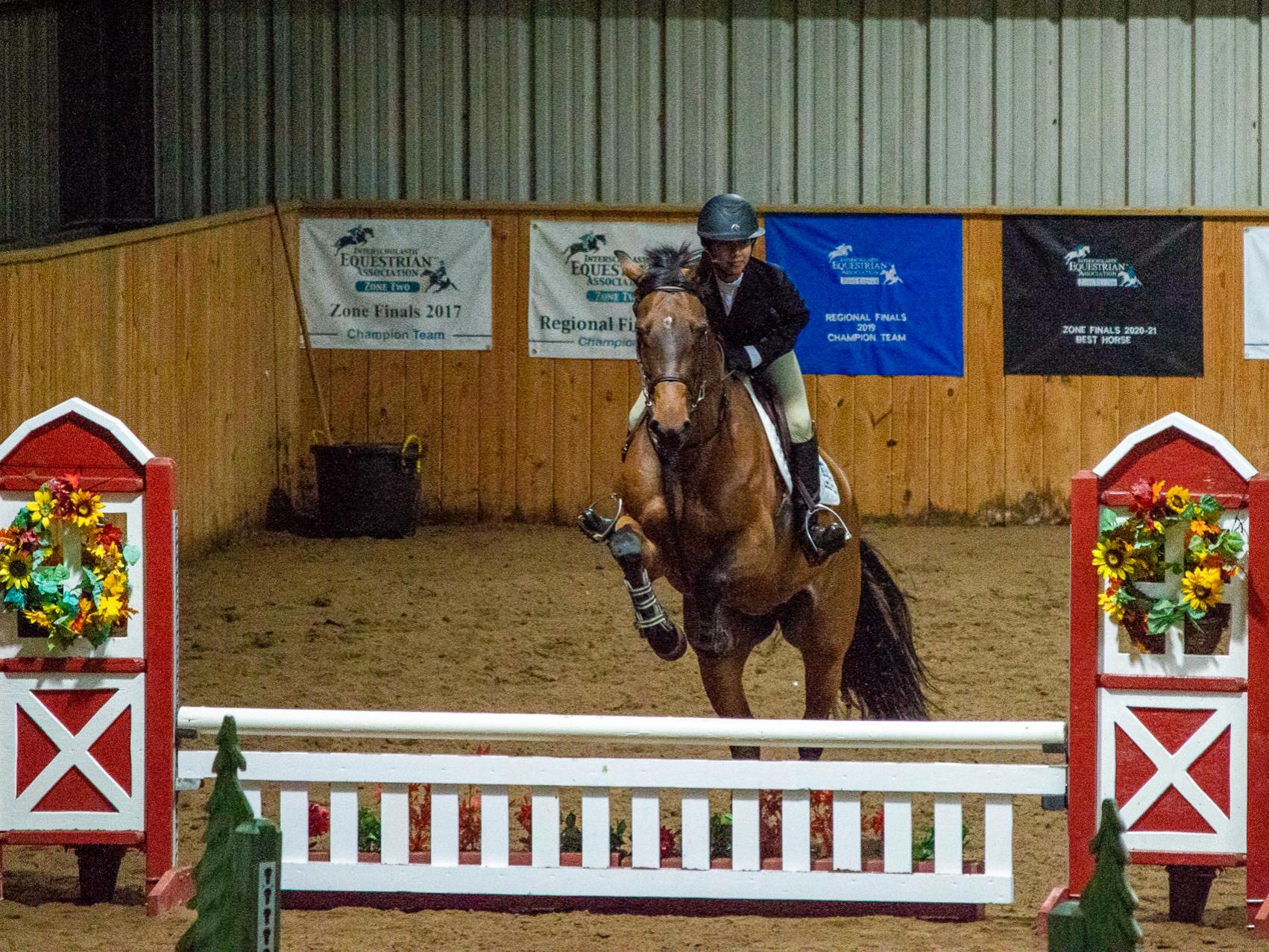 Jedily Alvarado on her horse while it jumps over a white fenced obstacle in a ring.