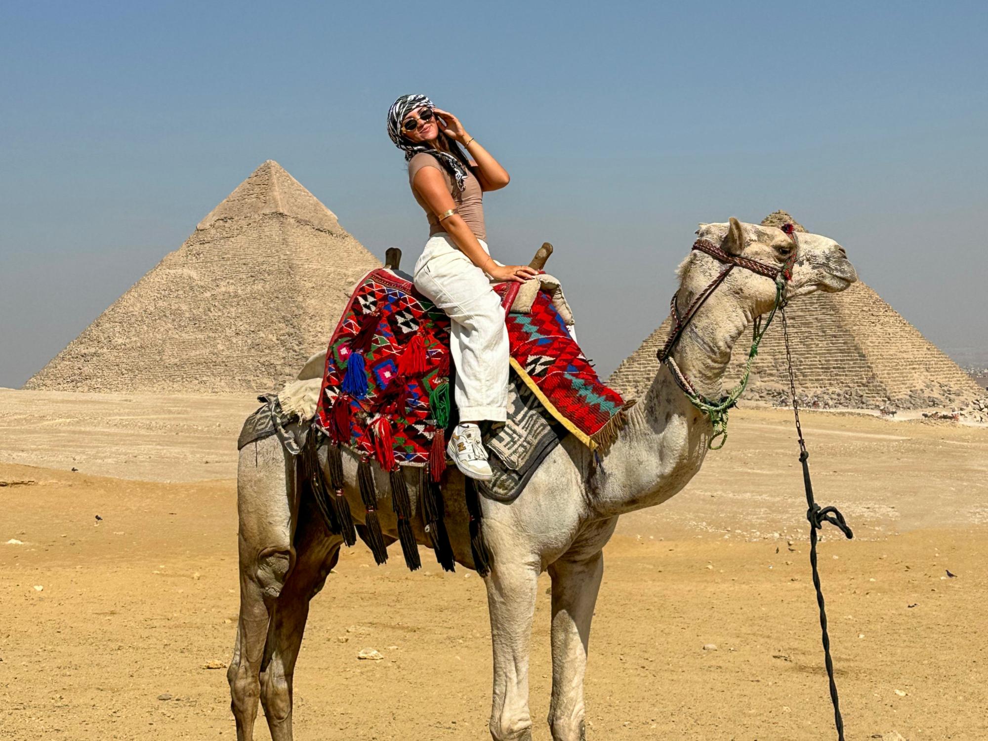 Lauren Fetterhoff sits on top of a camel in the dessert in front of two pyramids in Egypt.