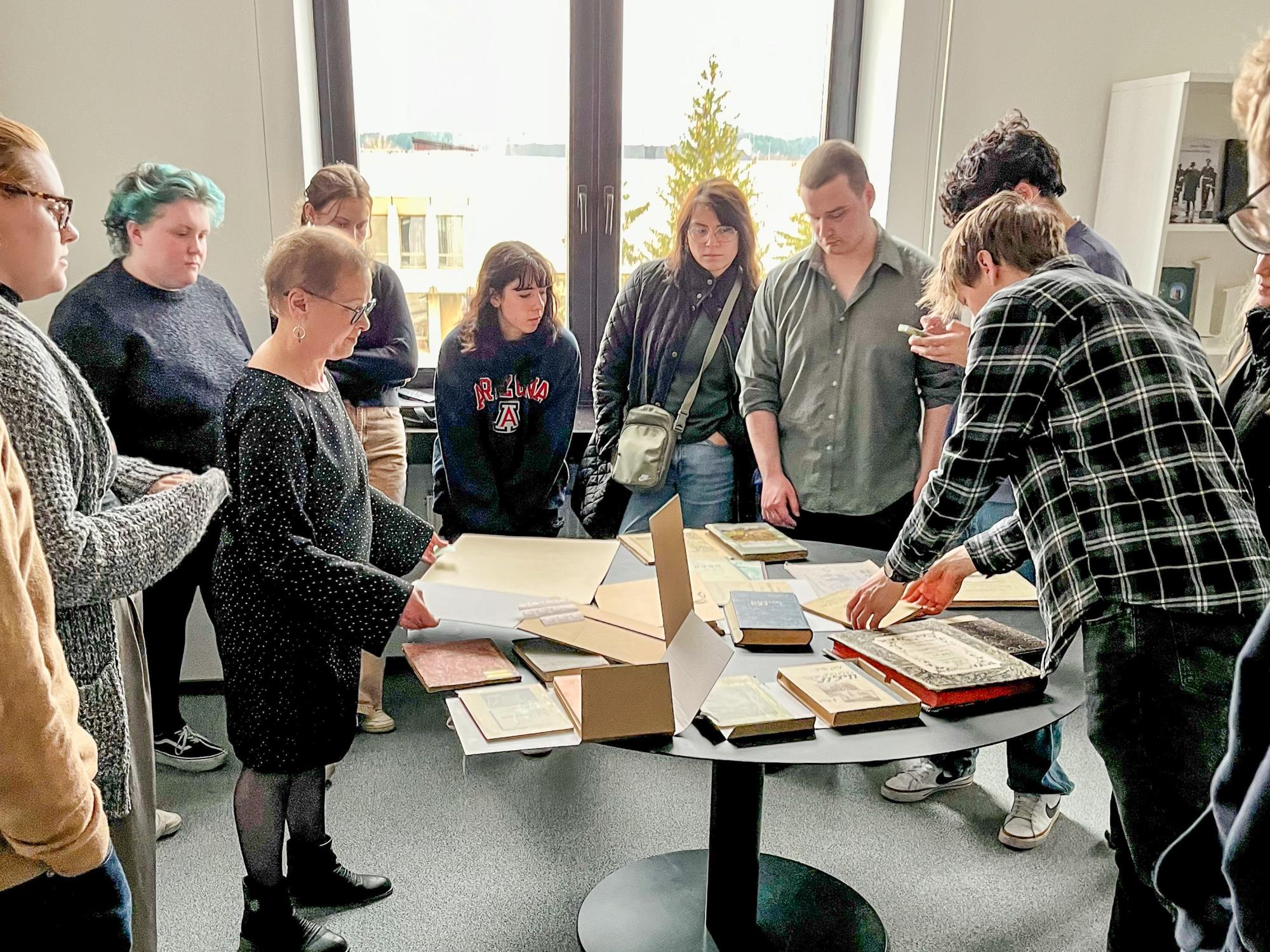 The class stands around a table covered in books in the Judaica section of the Lithuanian National Library with its director Lara Lempertiene.