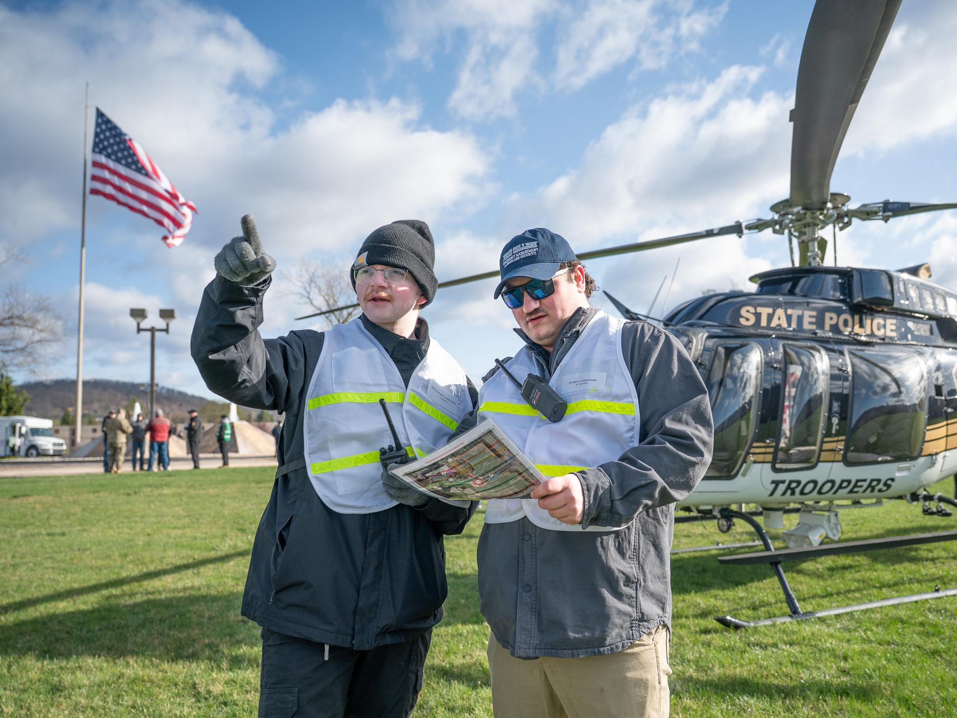 Two student wave at the camera with a helicopter behind them on the grass