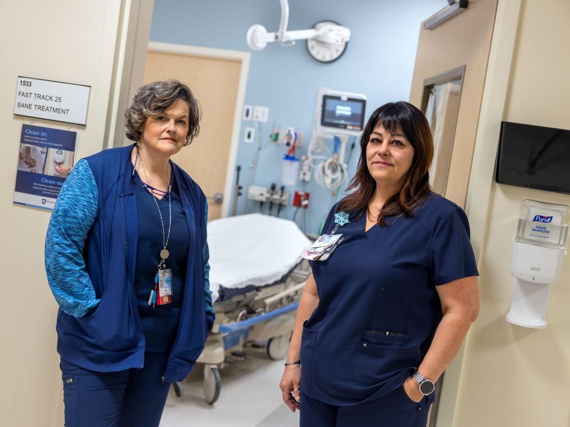Two nurses at Penn State Health St. Joseph Medical Center stand in the doorway of the Sexual Assault Nurse Examiner room. Vicky Reilly, left, is wearing a vest, top, pants and a name badge on a lanyard around her neck. Heather Owen, right, is wearing scrubs and a name badge pinned on the right of her top.