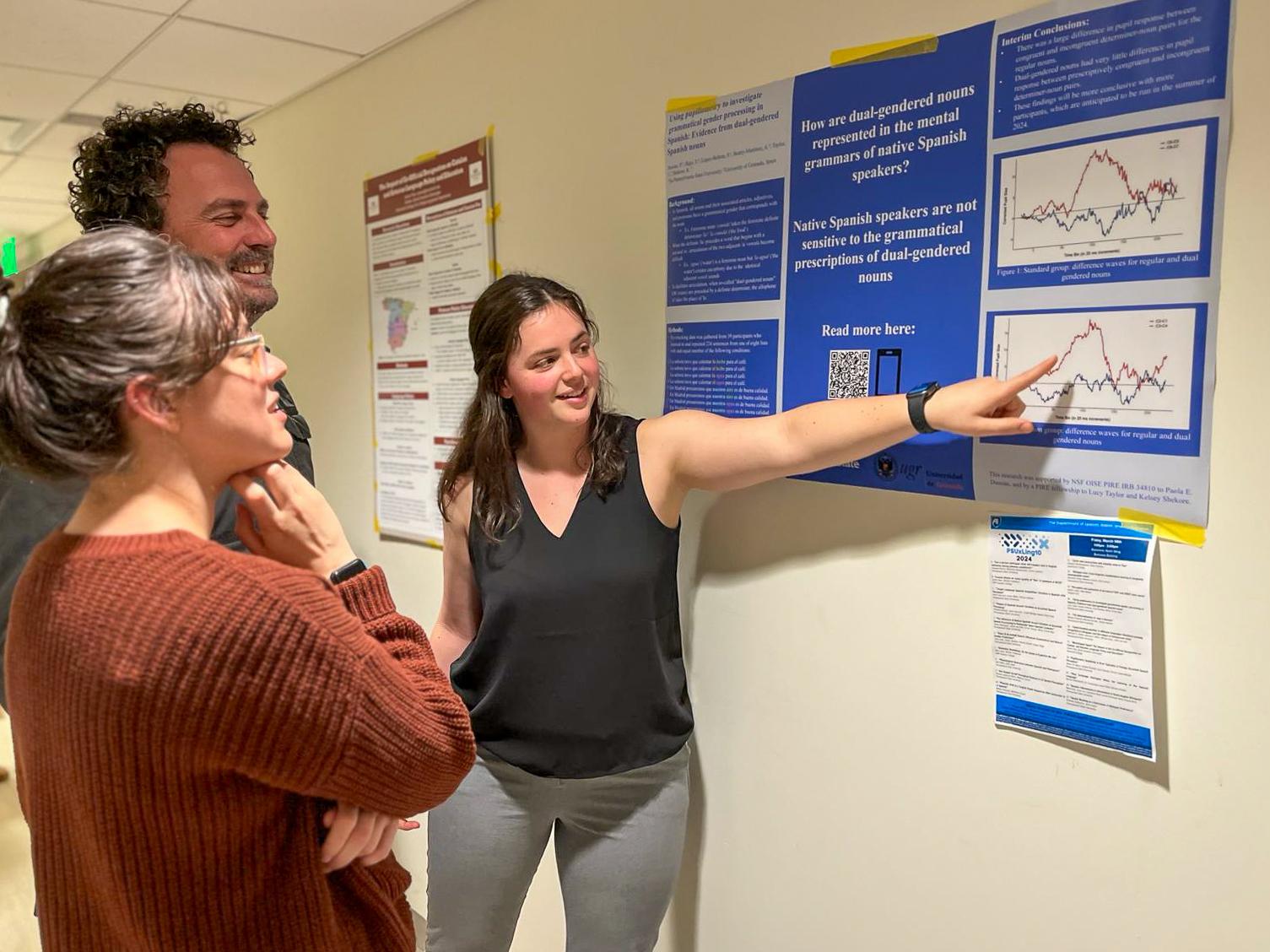 Lucy Taylor points to her PIRE research poster that hangs on the wall while two people listen intently.