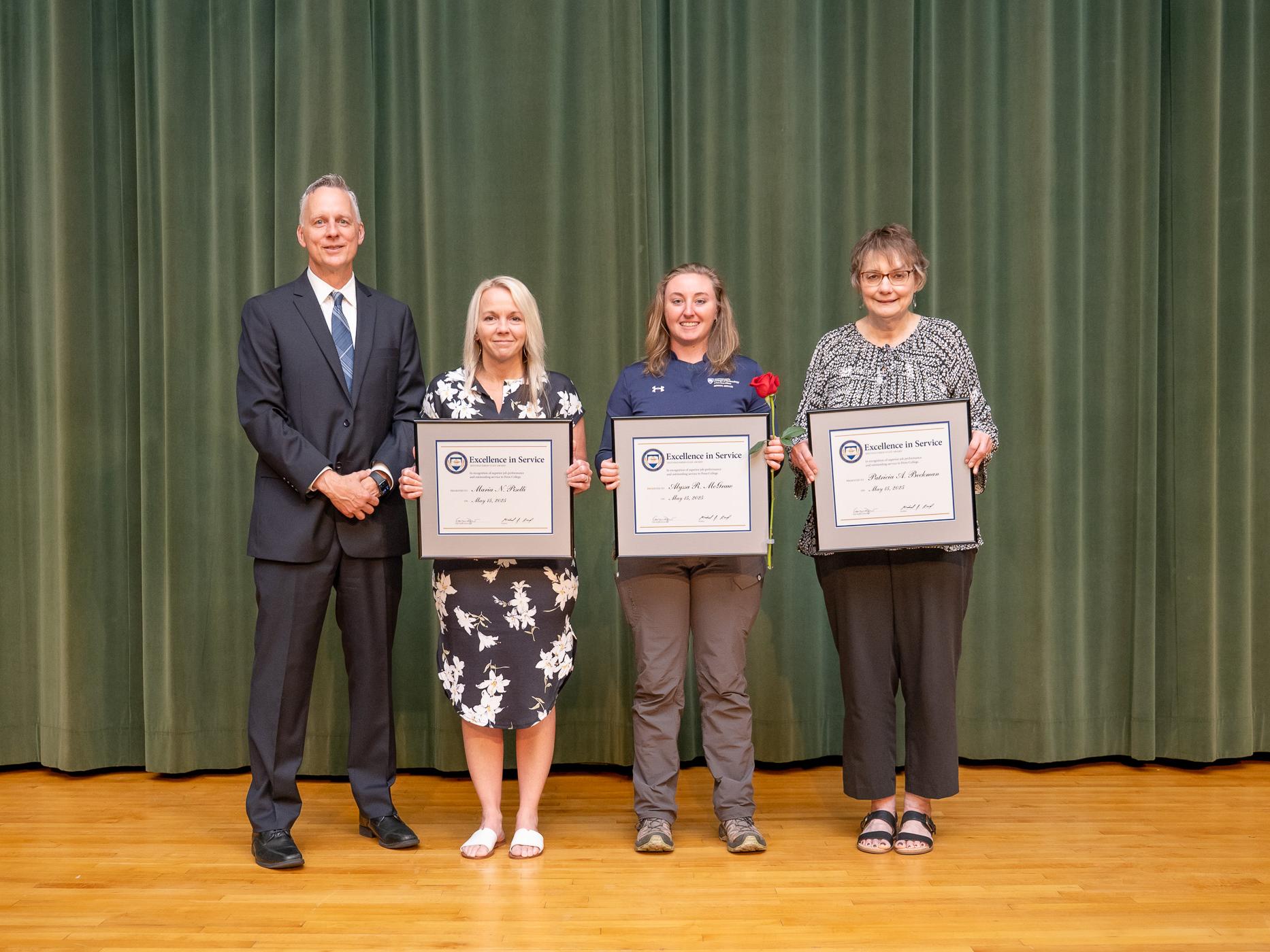 Four people pose on an auditorium stage, with three holding framed awards