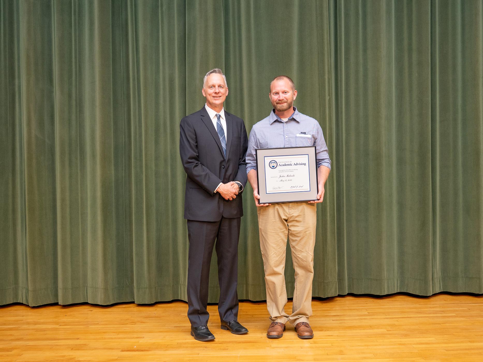 Two people pose together on a stage, one holds a framed award
