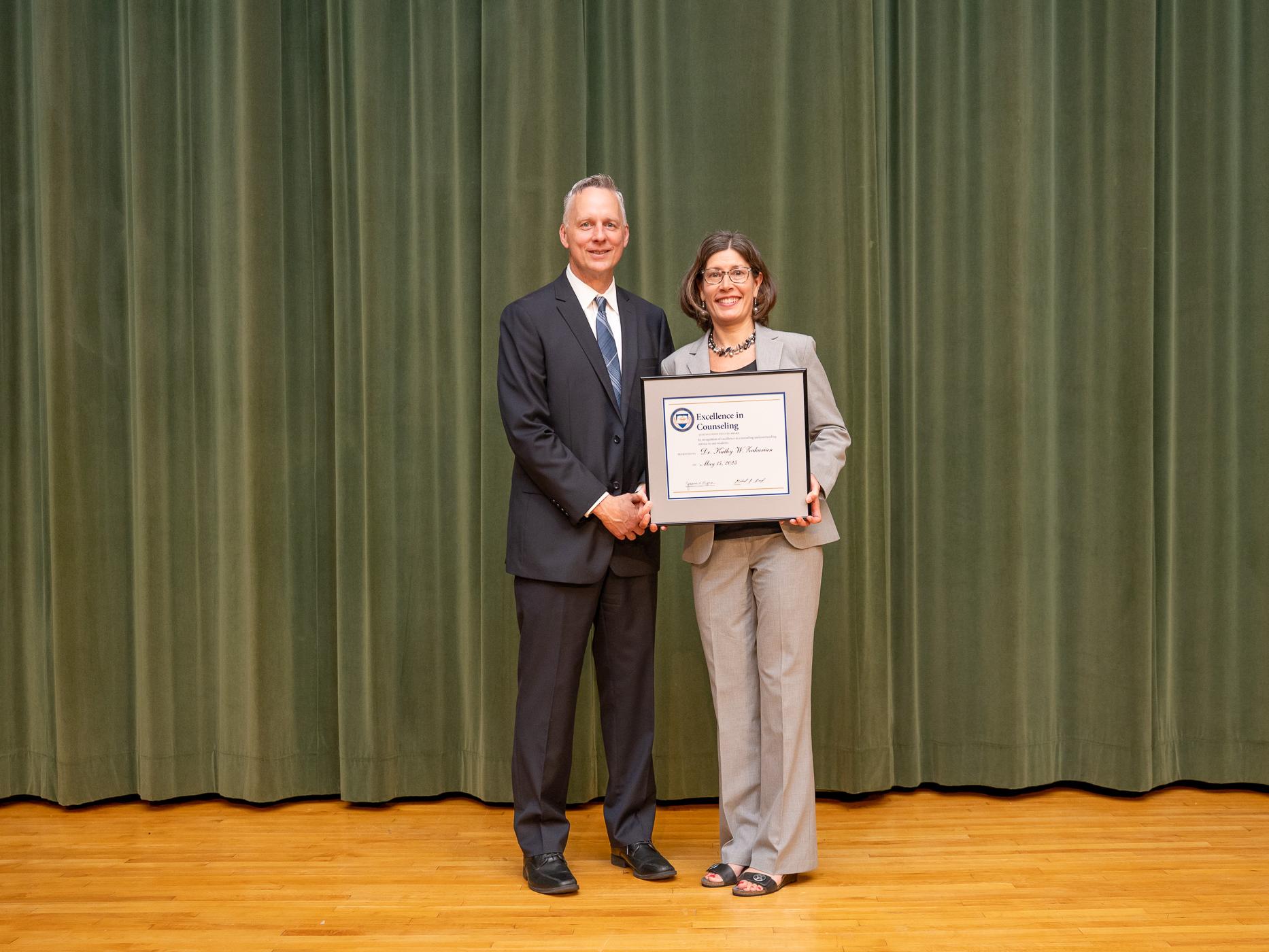 Two people pose on a stage, one holds a framed award