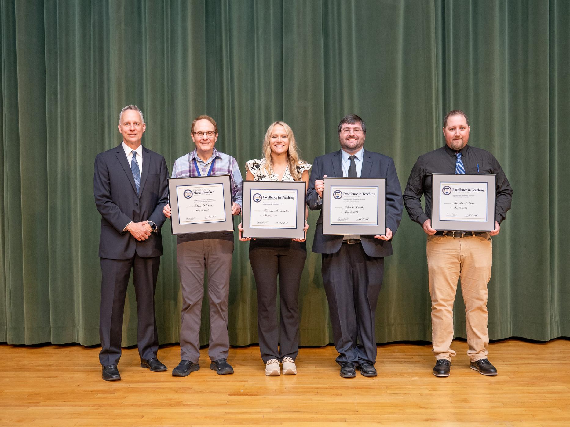 Five people pose for a photo in an auditorium, with four of them holding framed awards