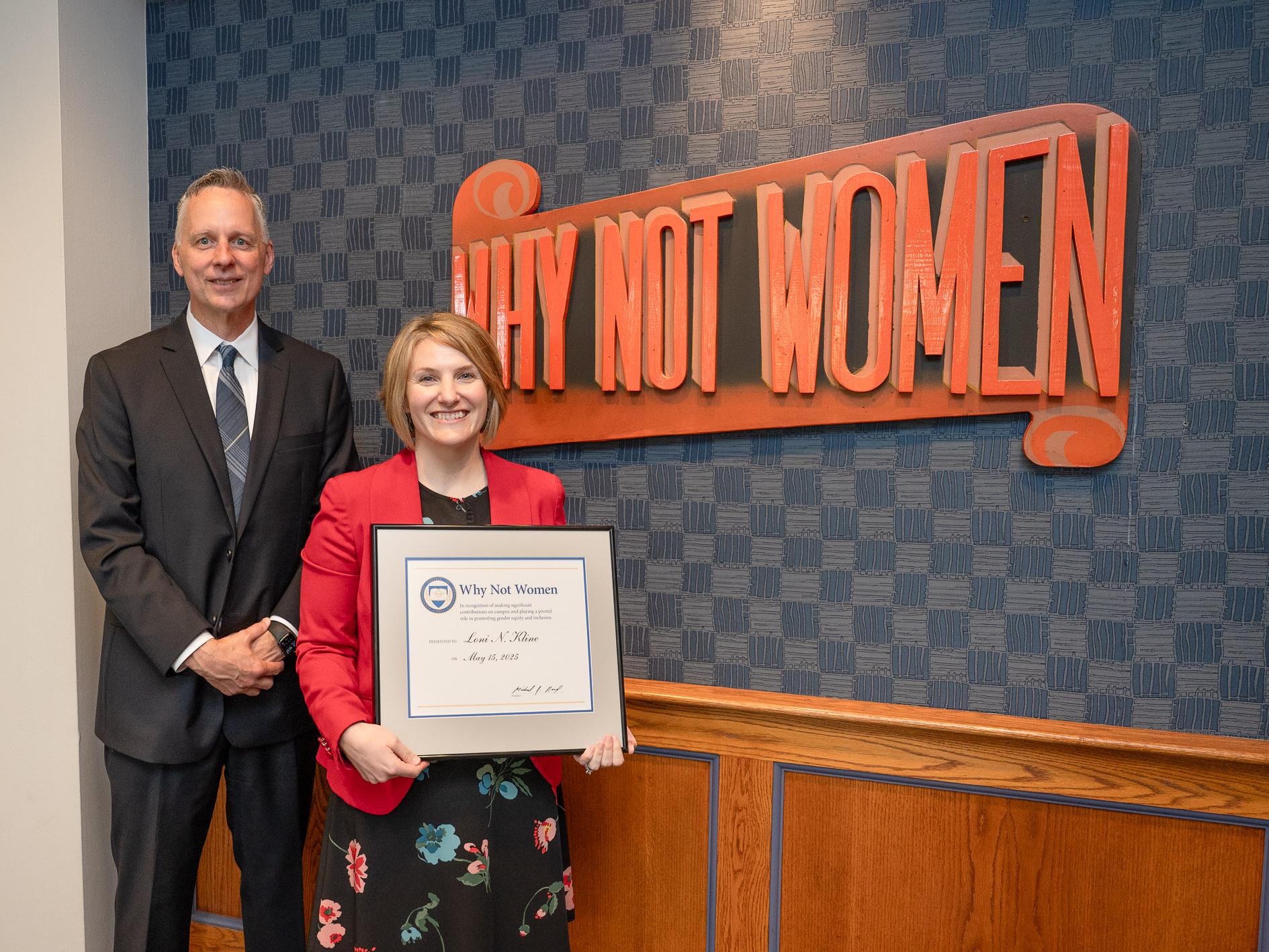 Two people pose on a stage, one holds a framed award