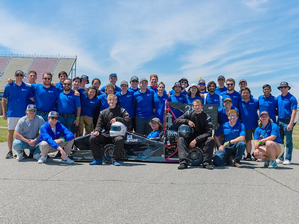 Students wearing blue shirts pose outdoors with a racecar on a racetrack. 