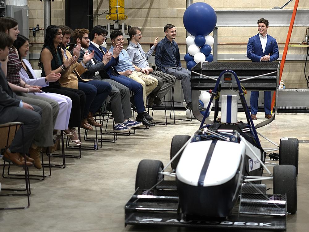 Students clap as a racecar is unveiled in a large room with blue and white balloons.  