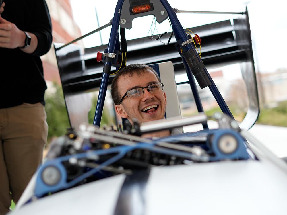 An individual wearing glasses smiles while seated in a racecar.  