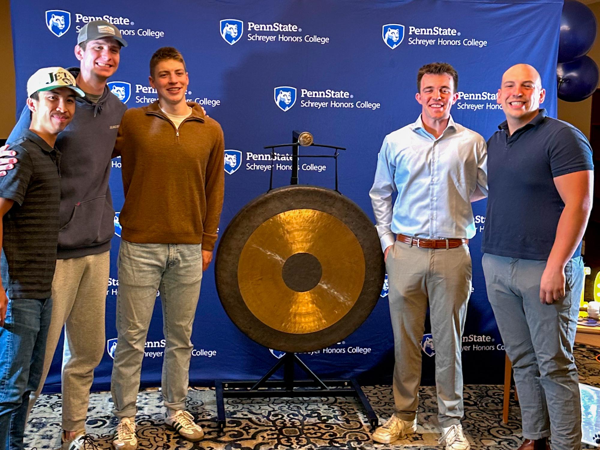 Five men stand in front of a blue backdrop with the Schreyer Honors College wordmark with a gold gong in the middle. 