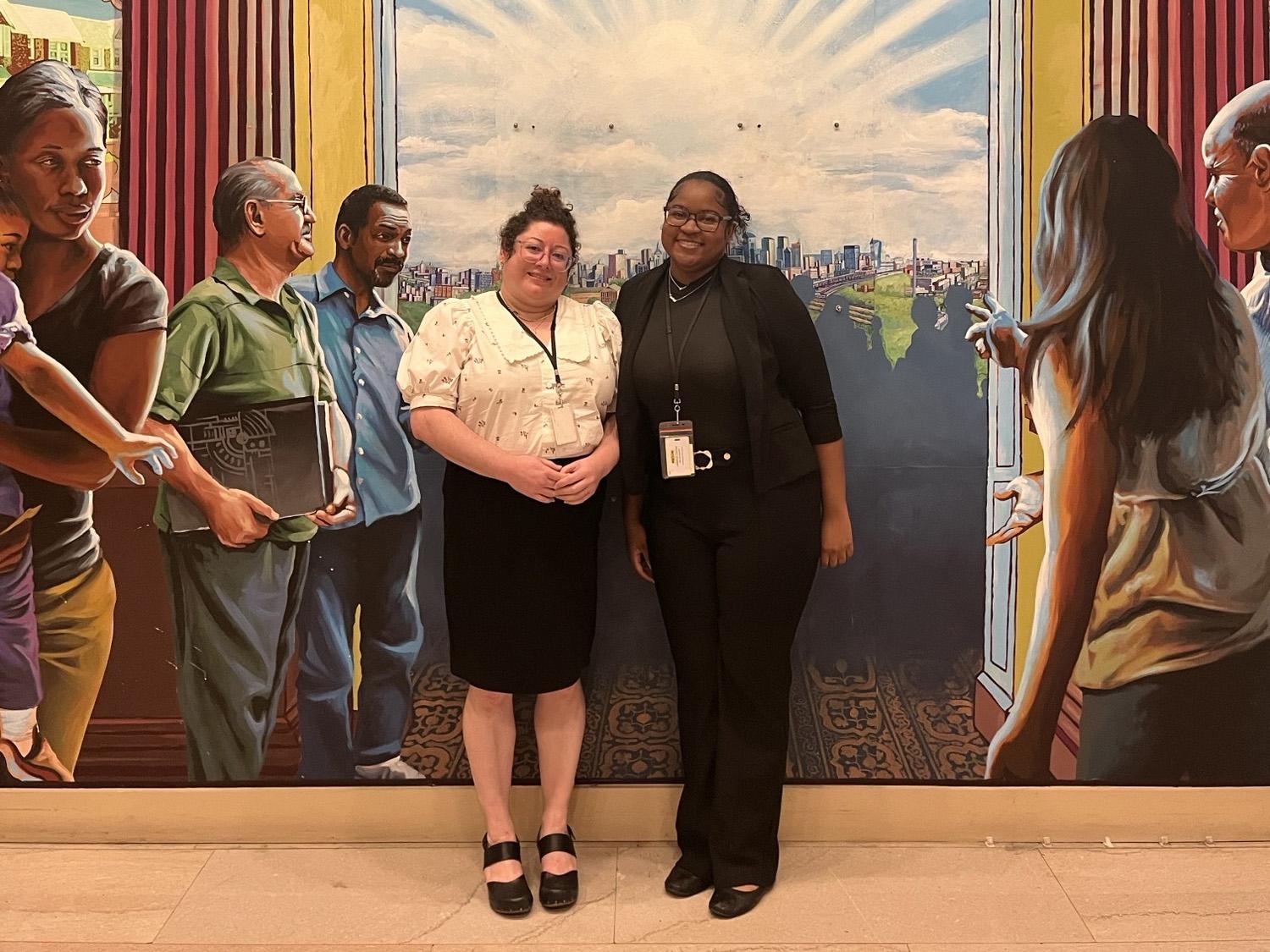 Cameryn Allen (right) and her supervisor Bethany Watzman (left) stand in front of a mural in the lobby of the Philadelphia District Attorney’s Office.