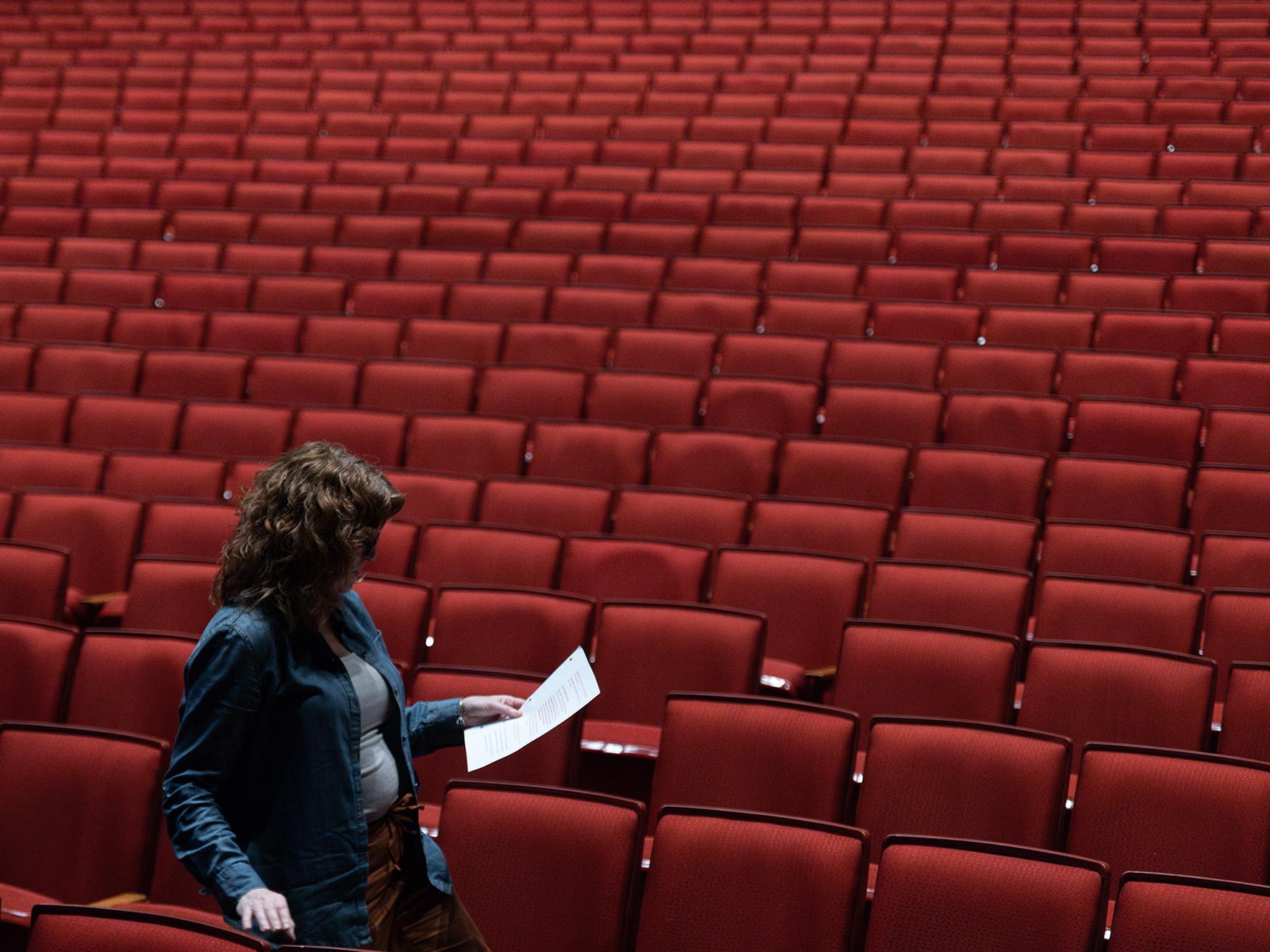 A woman looks at a sheet of paper and walks through an auditorium of empty red seats