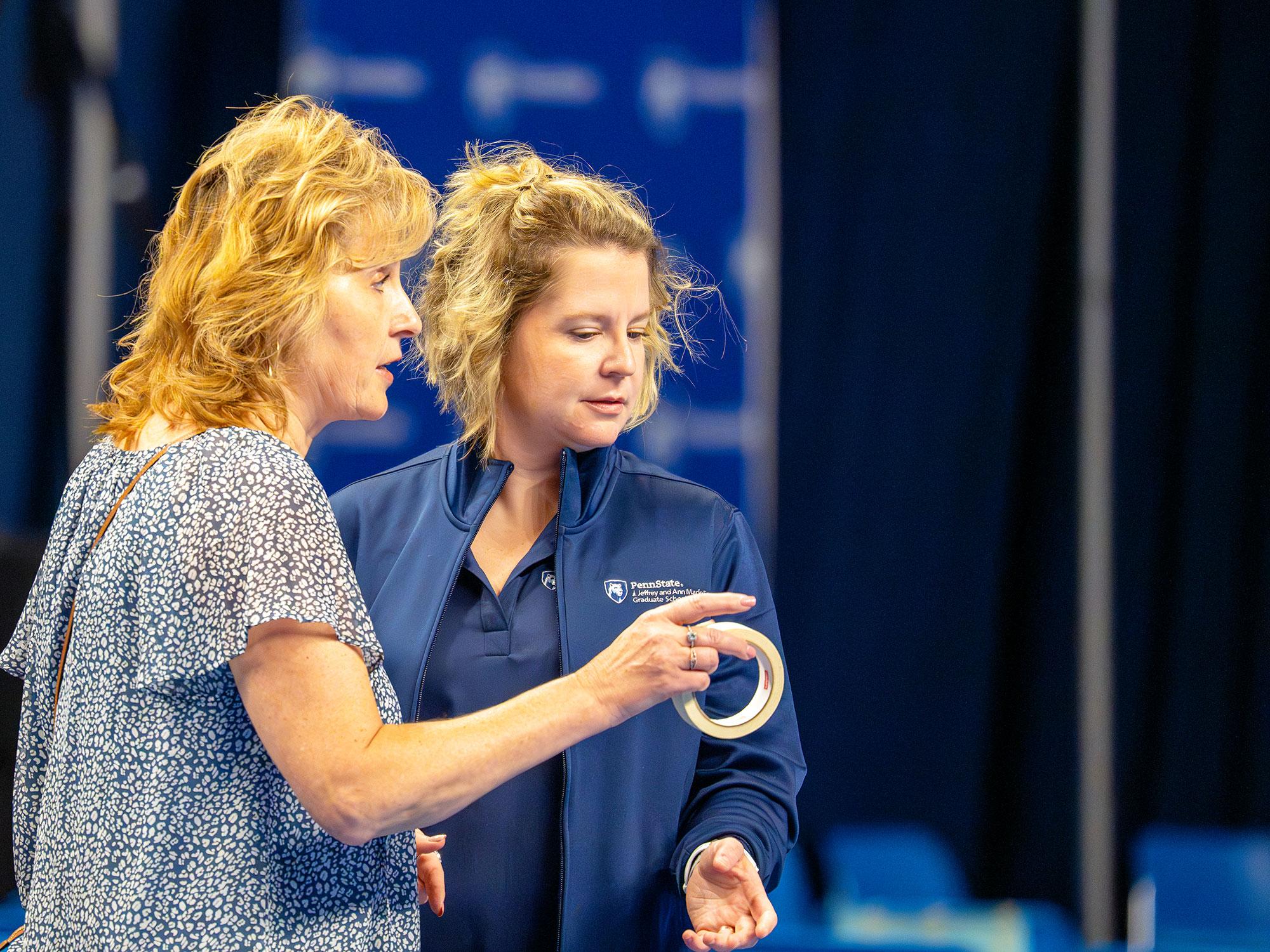 Two women speak to each other in front of rows of blue plastic chairs