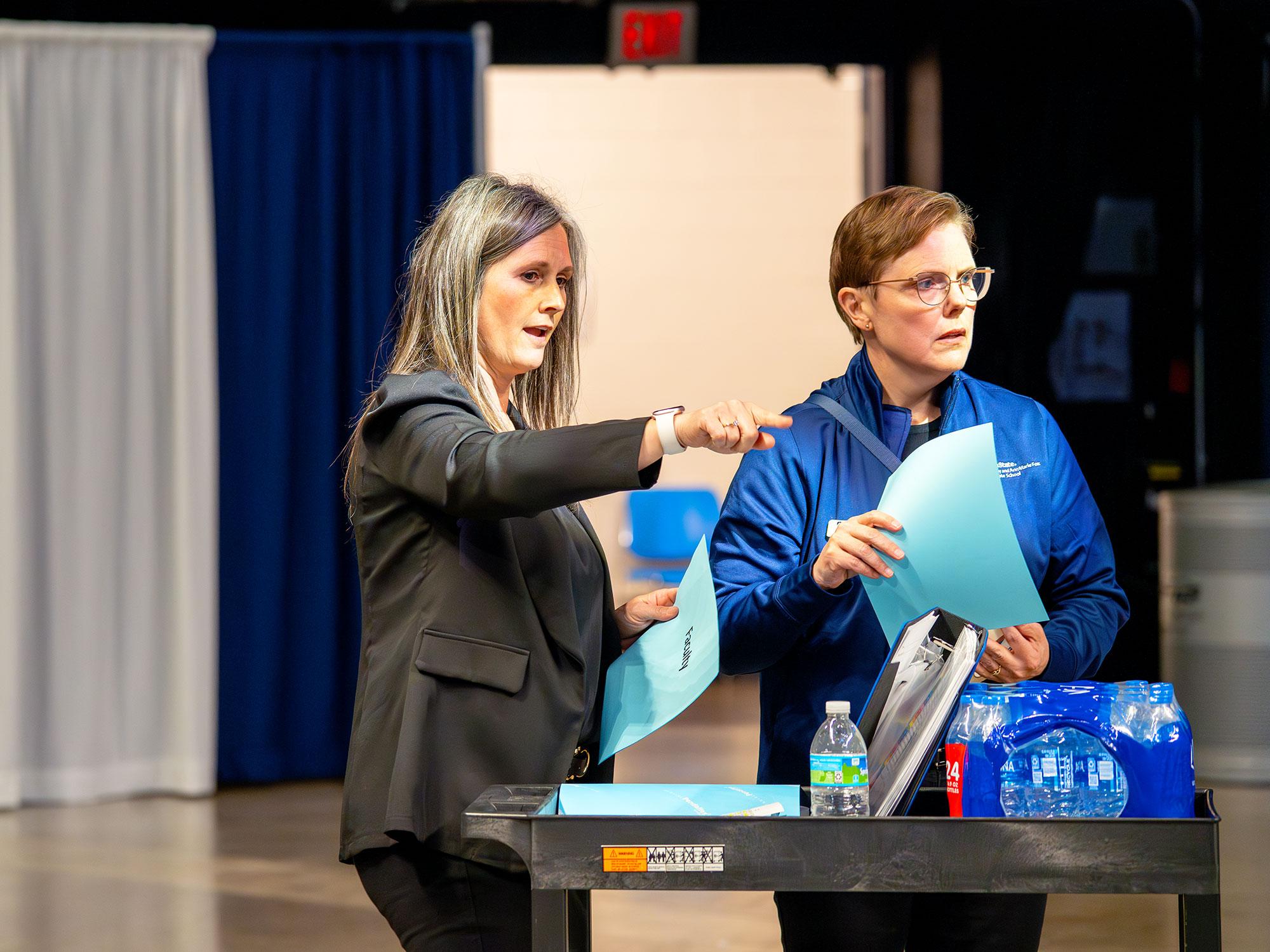 Two women stand in an arena and gesture toward the space in front of them