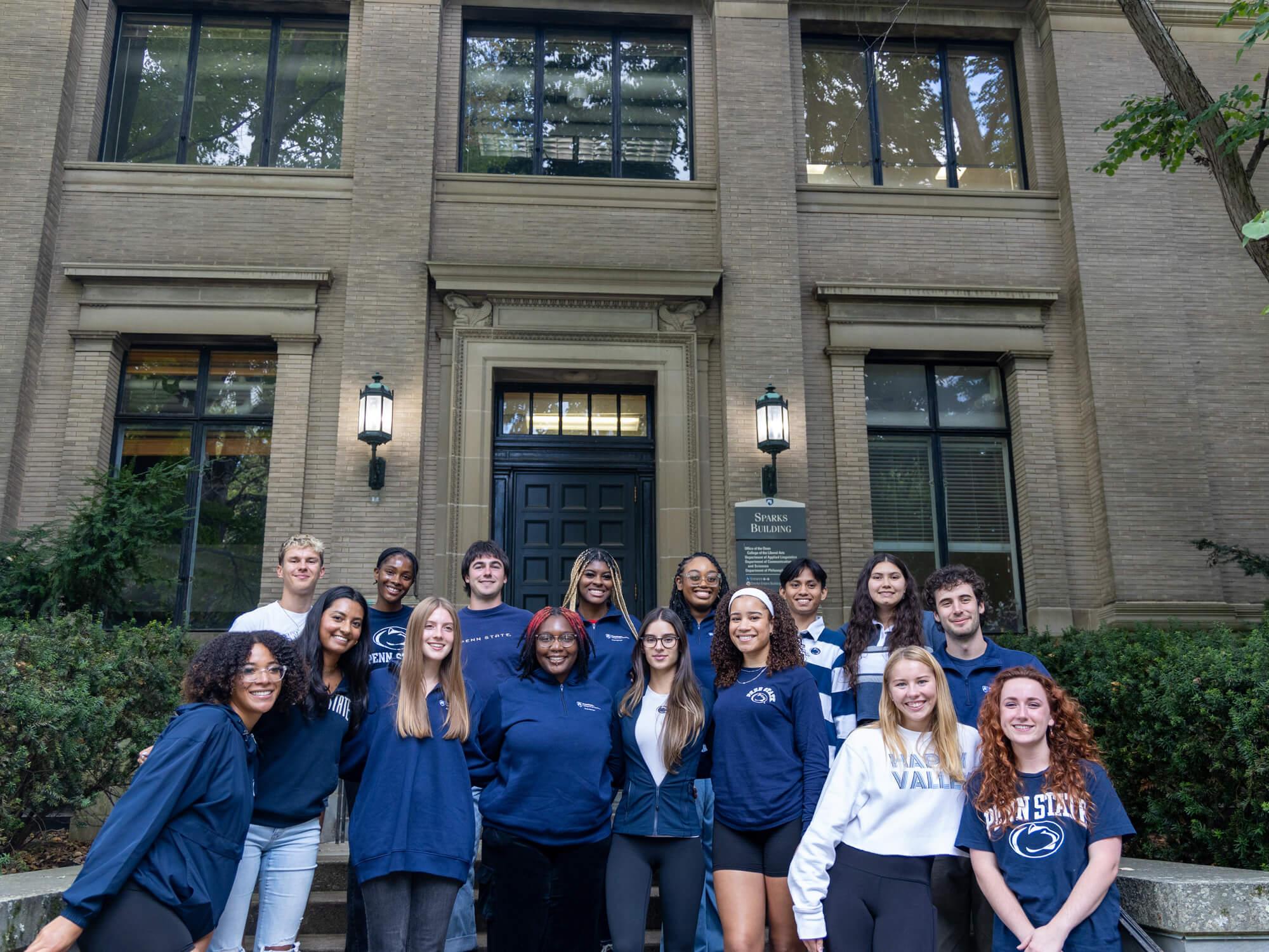 A group of students smiling in blue and white Penn State attire in front of Sparks Building.