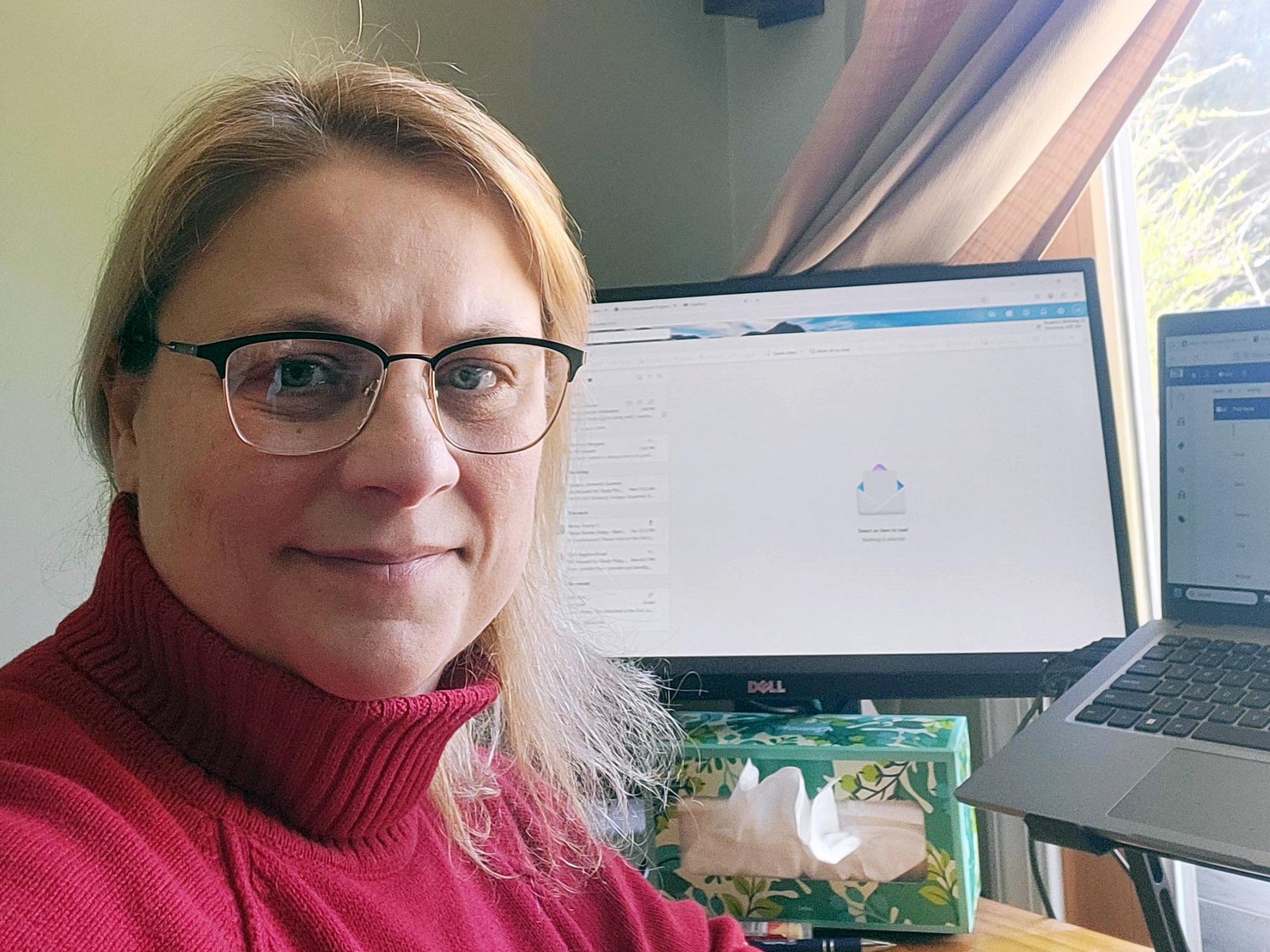 A woman sits in front of a computer monitor in a home office and smiles at the camera.