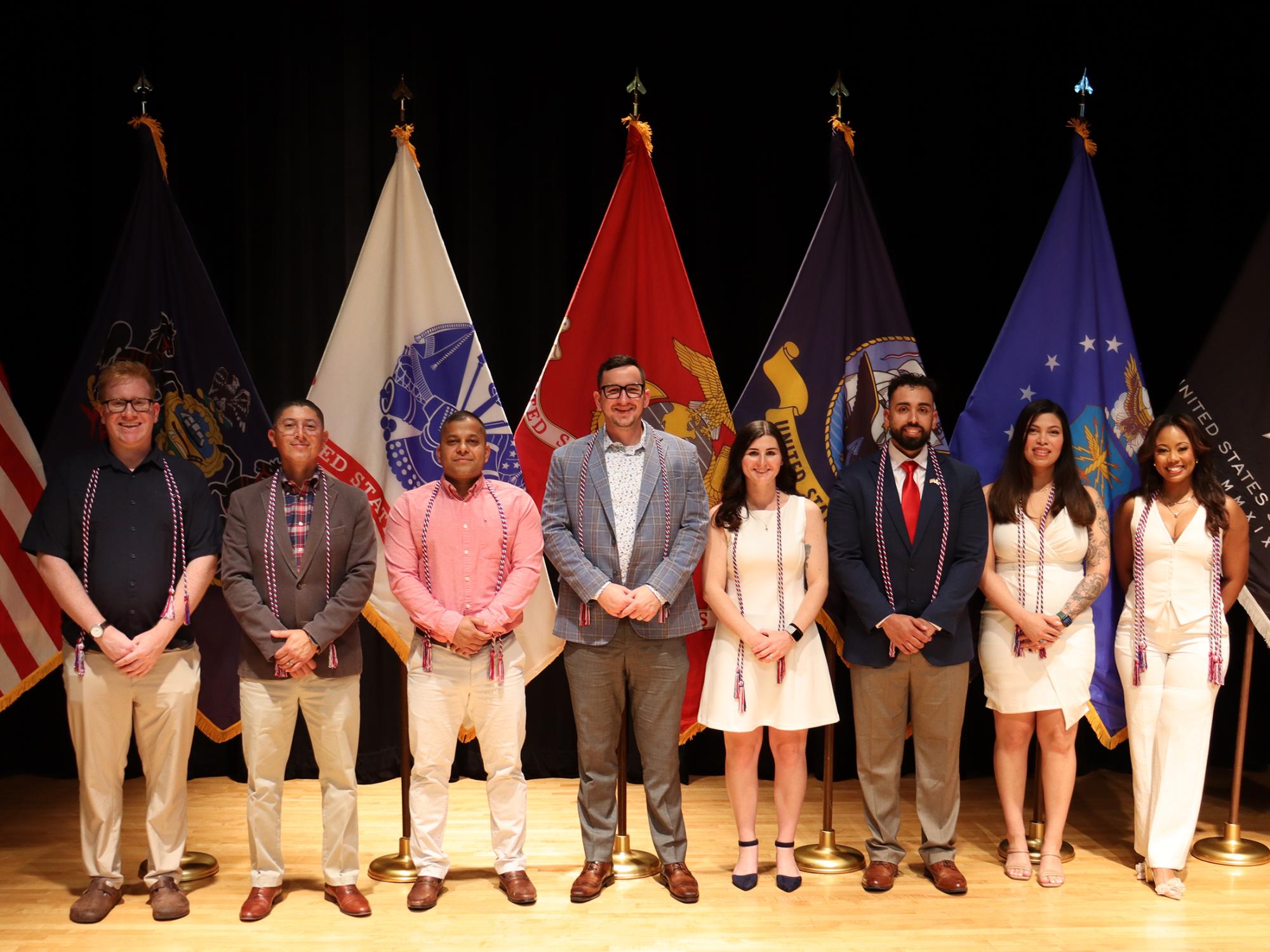 A group of people lined up in front of U.S. military flags, the American Flag, and the Pennsylvania flag.