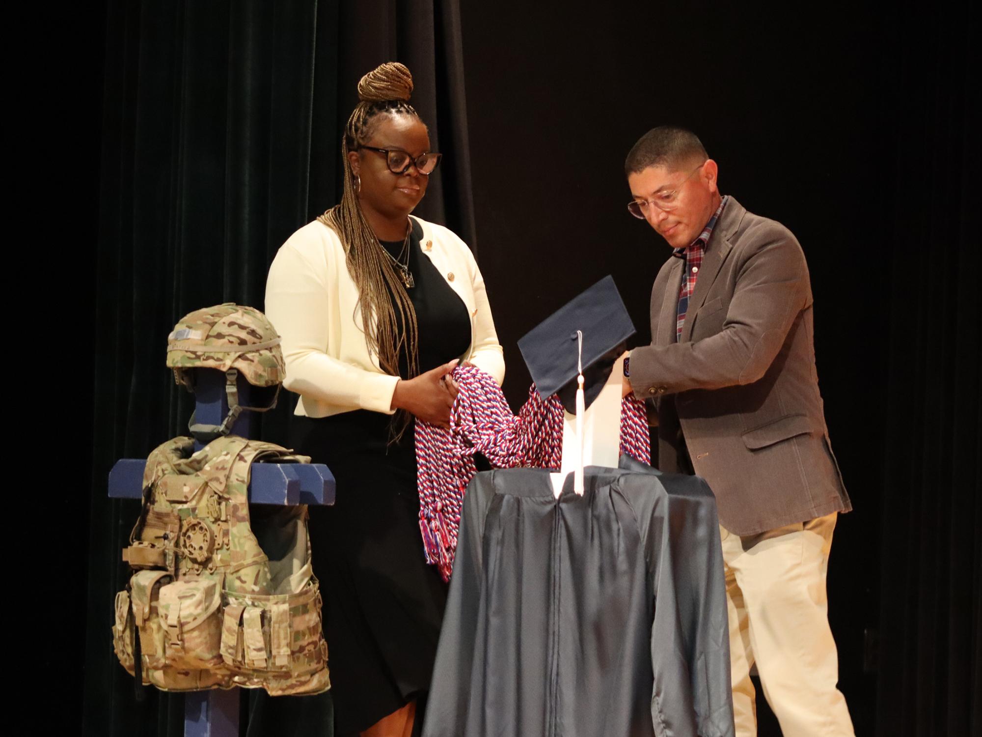A woman and a man together lift a bundle of red, white, and blue honor cords on a stage.