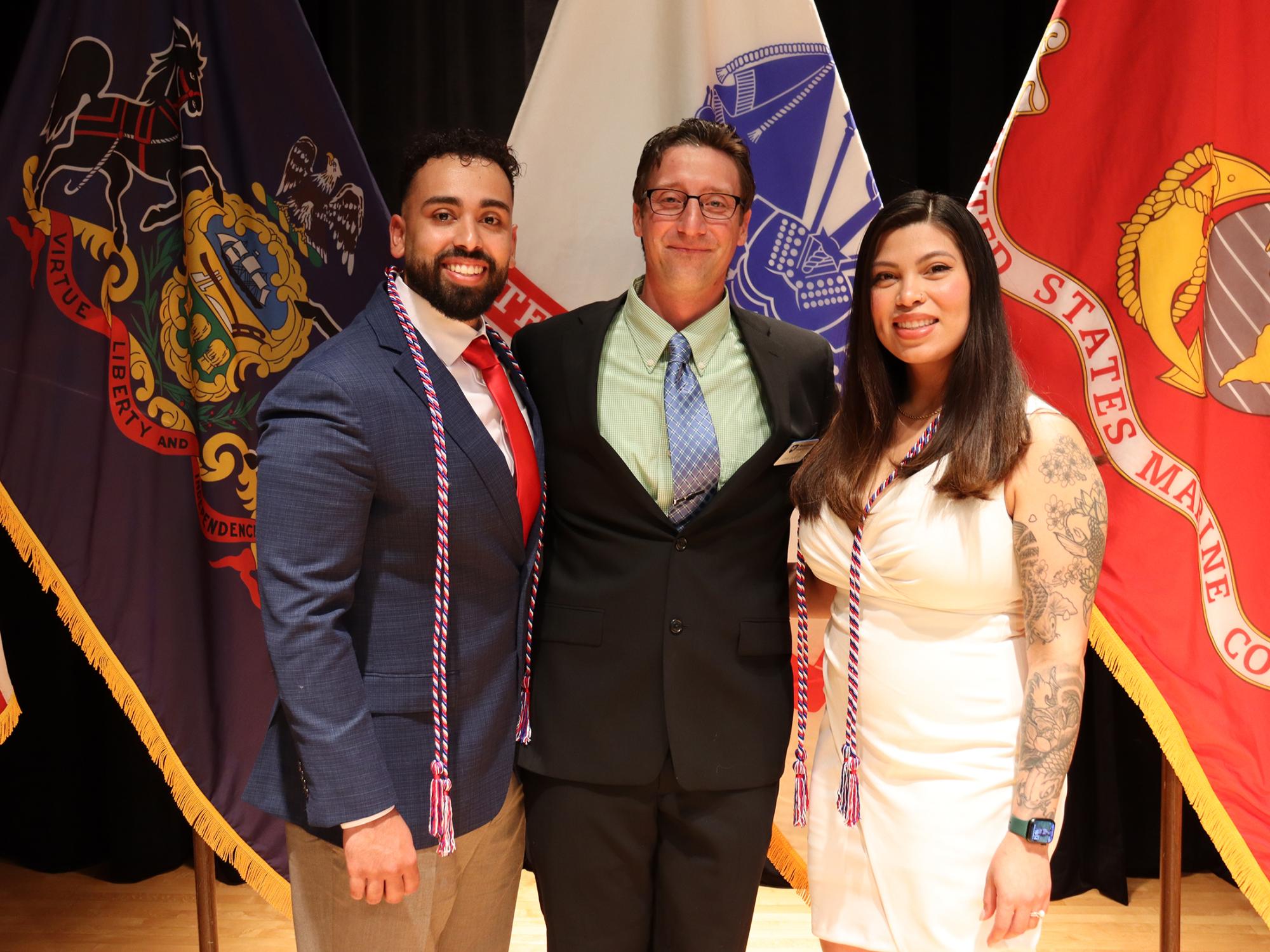 Two men wearing suits and a woman in a white dress pose in front of a group of flags.