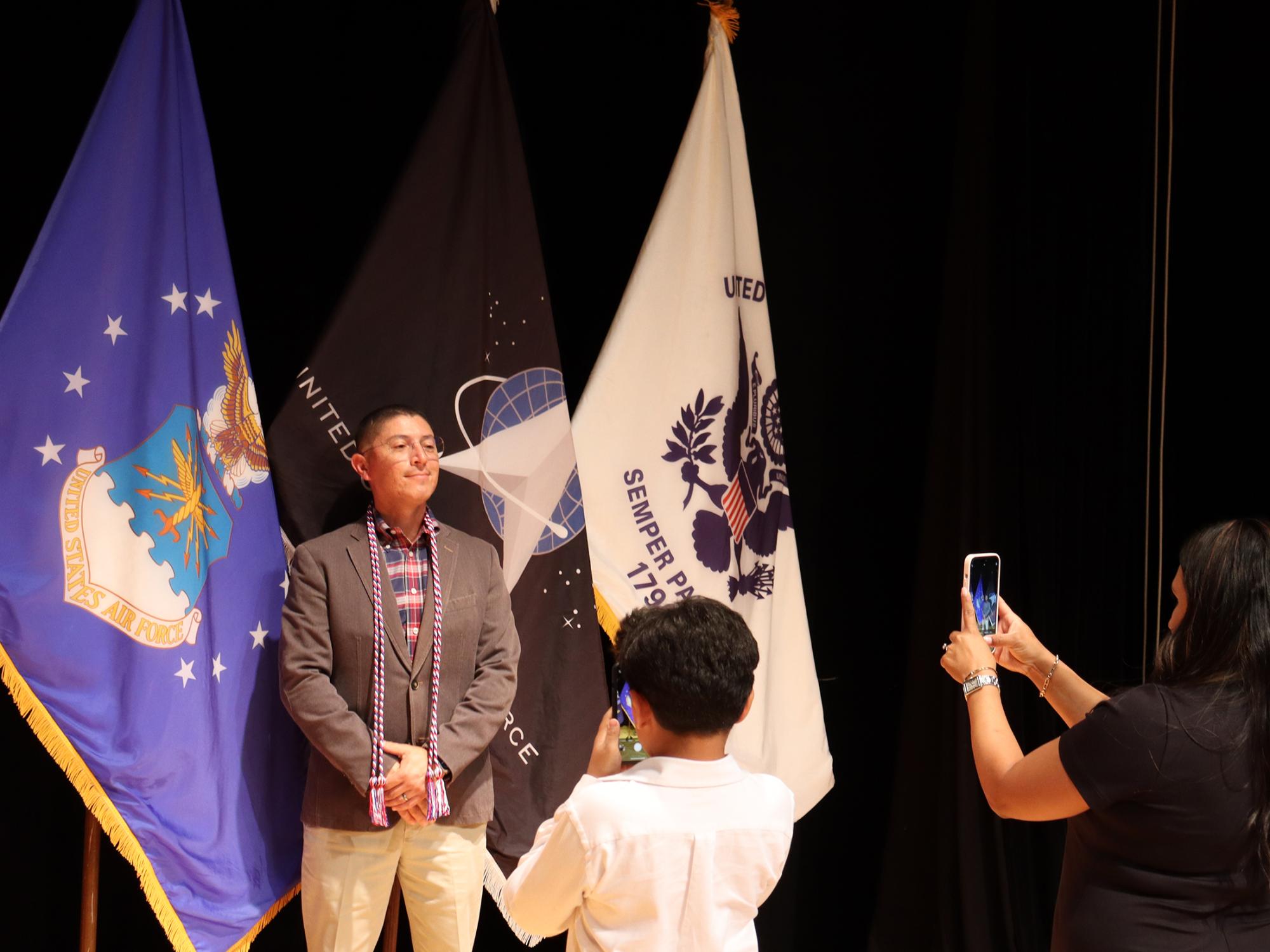 Two people take a photo of a man posing in front of U.S. military flags.