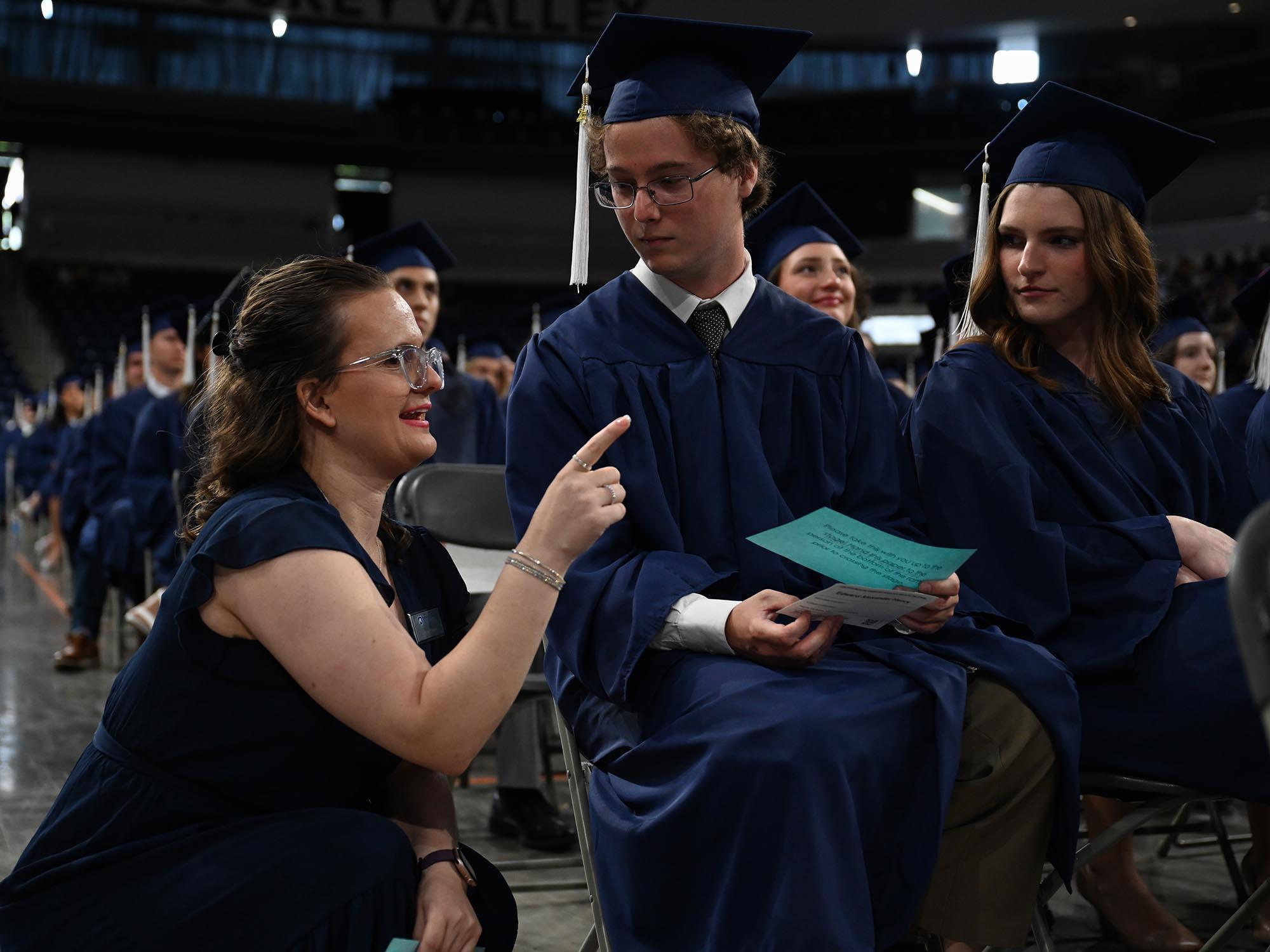 A woman kneels next to a student in graduation ceremony attire and speaks to him, pointing out of frame.