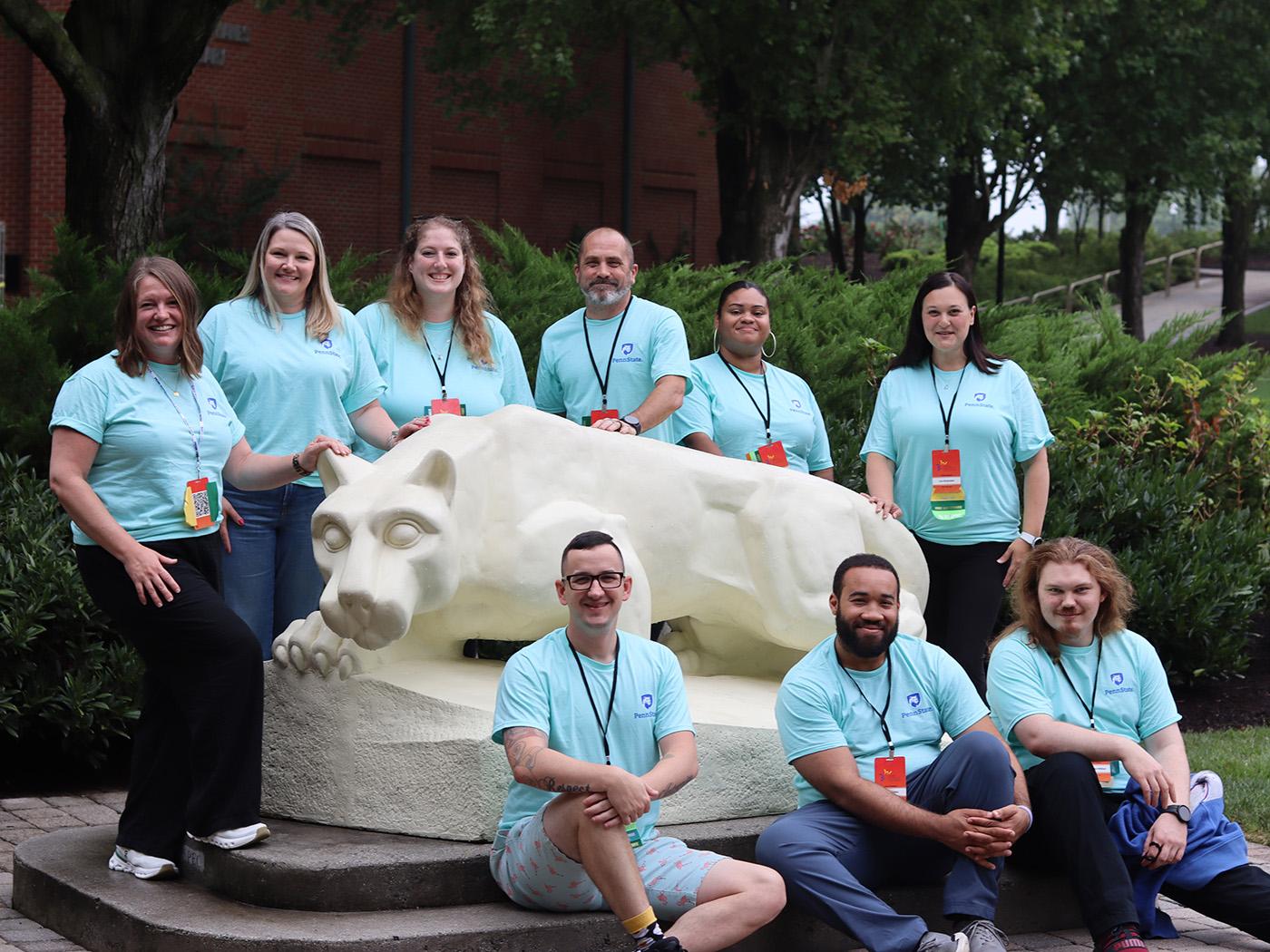 A group of people wearing aqua blue T-shirts pose around the Nittany Lion Shrine at Penn State Harrisburg.
