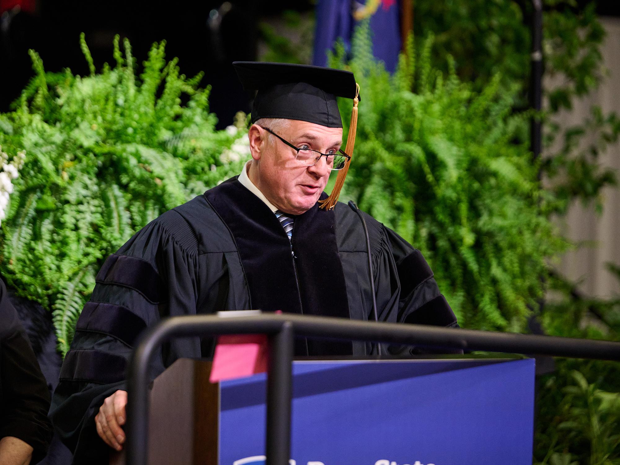 A man in commencement regalia stands at a podium and looks out over the crowd