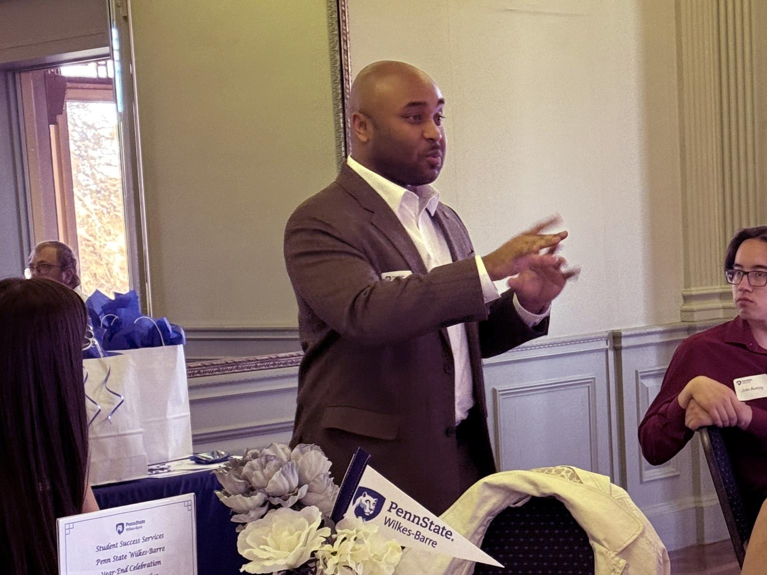 A man standing aside a table gestures while speaking.