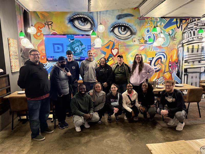 A group of students pose for a photo in front of a mural.