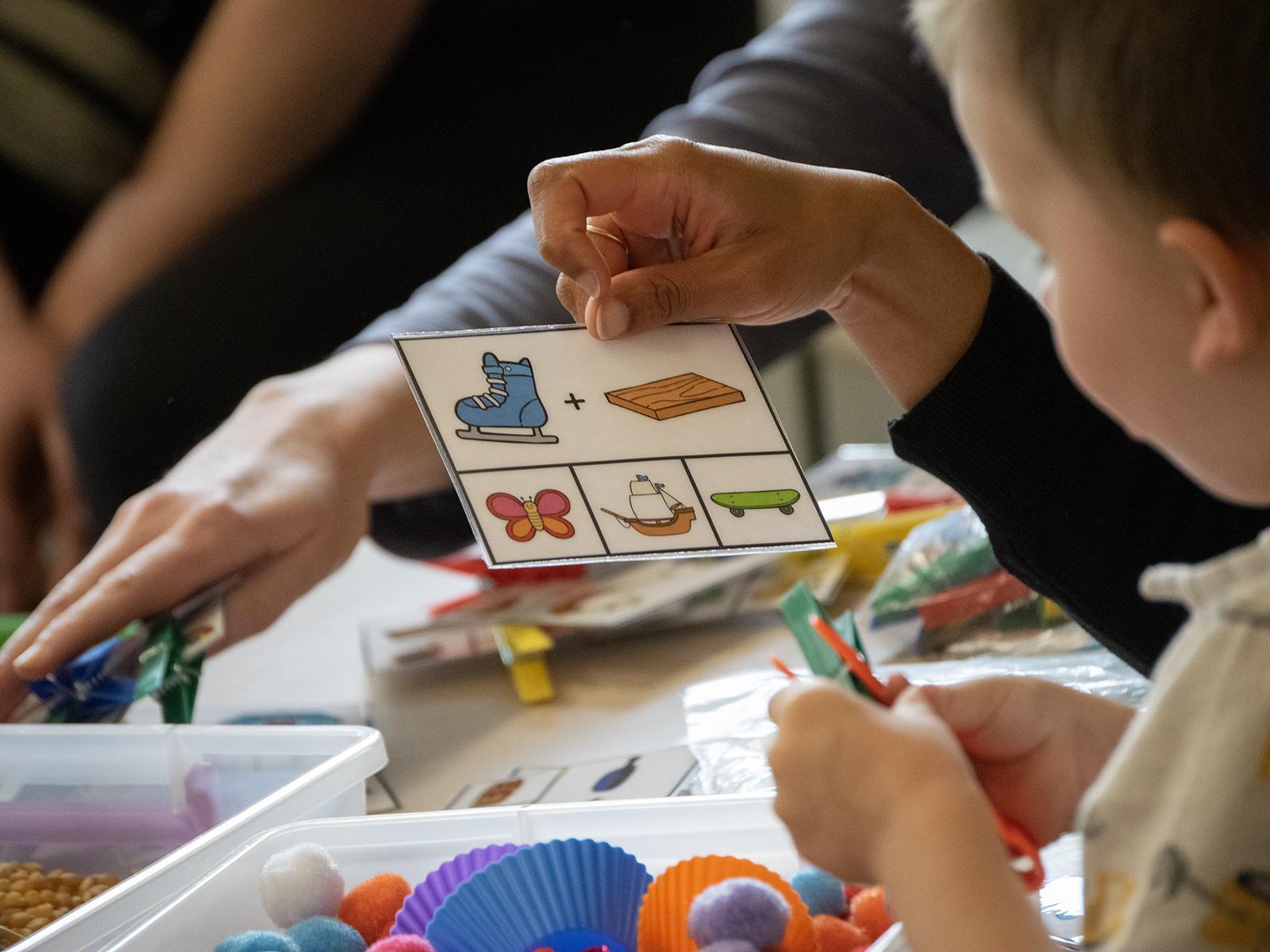 A child looks at symbols on a card, with a sensory bin of cotton balsl in front of him