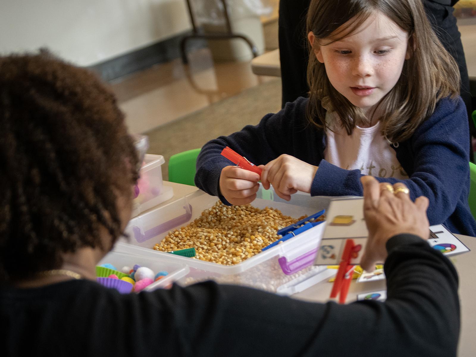 A child looks at symbols on a card, with a sensory bin in front of her