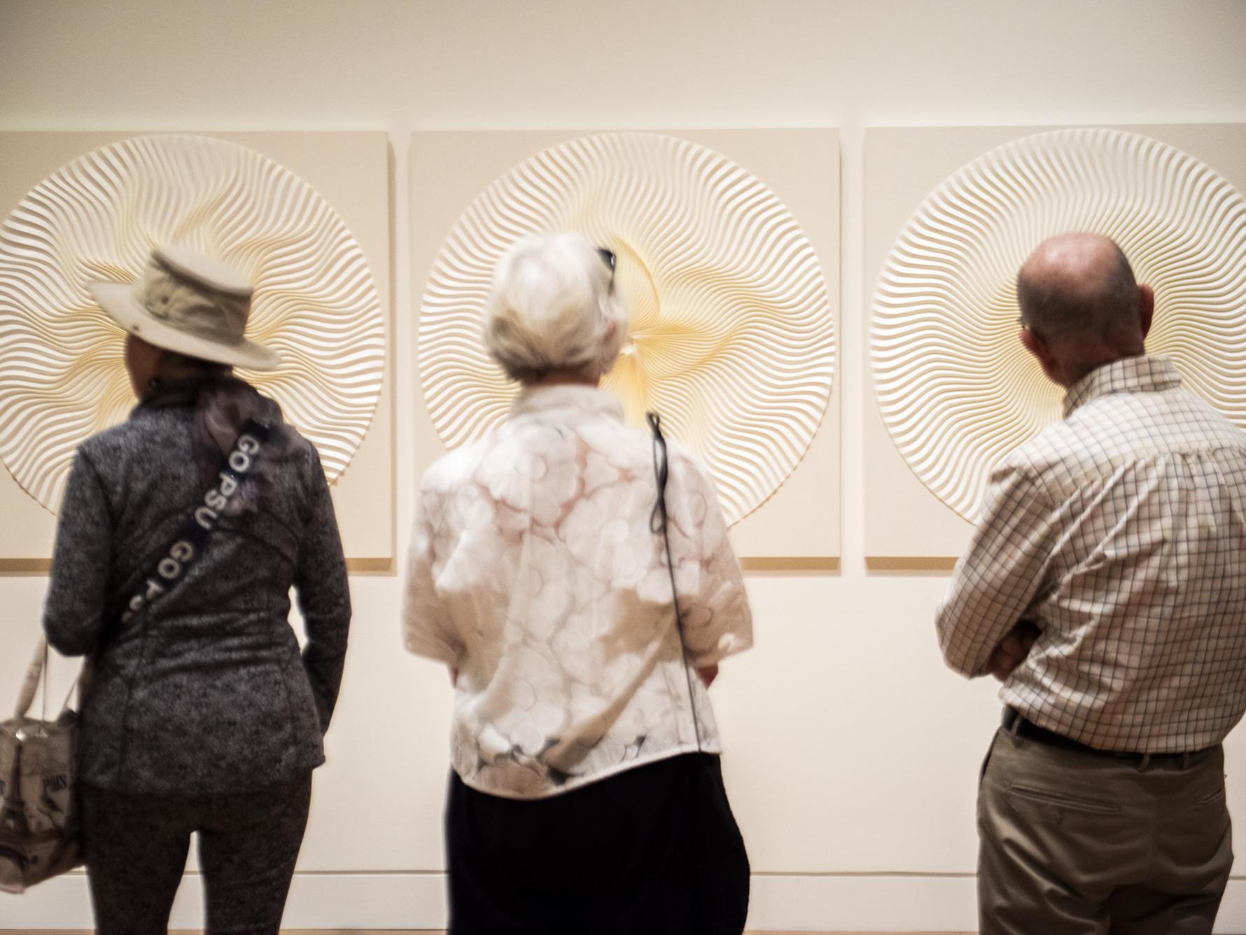 Three people in front of three works of white folded paper