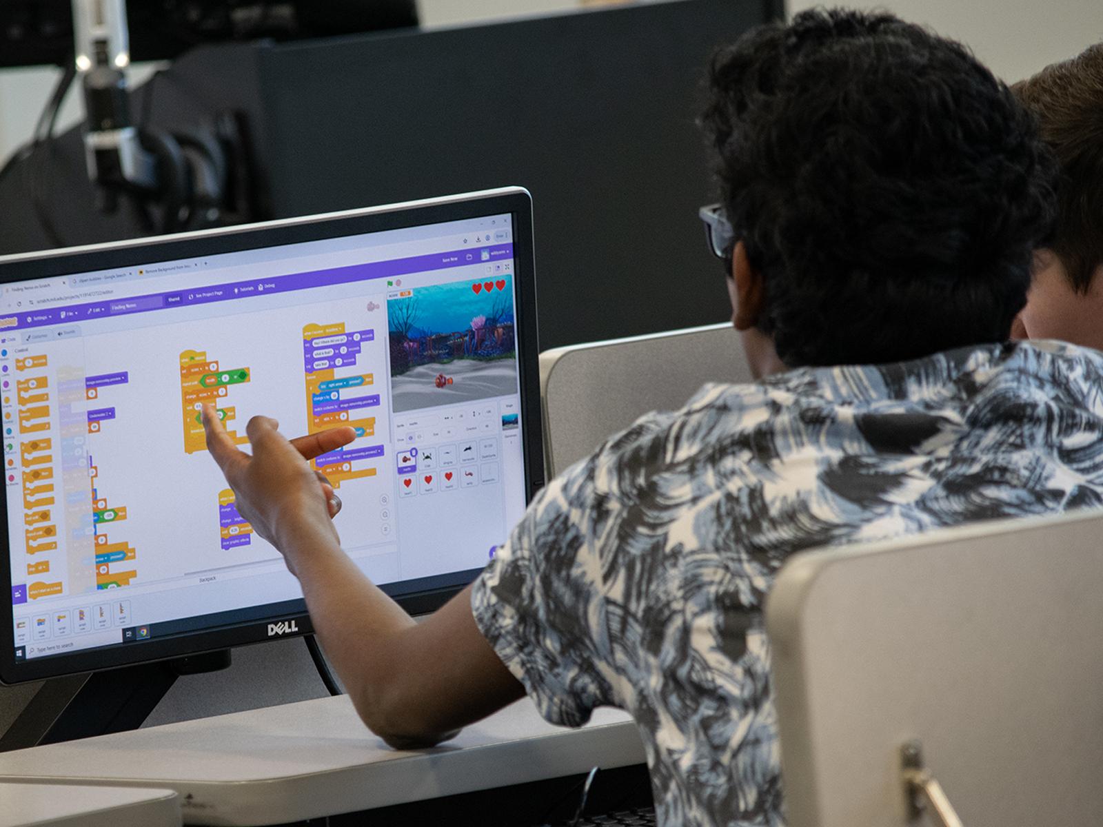 A student points to a computer screen showing a colorful coding activity