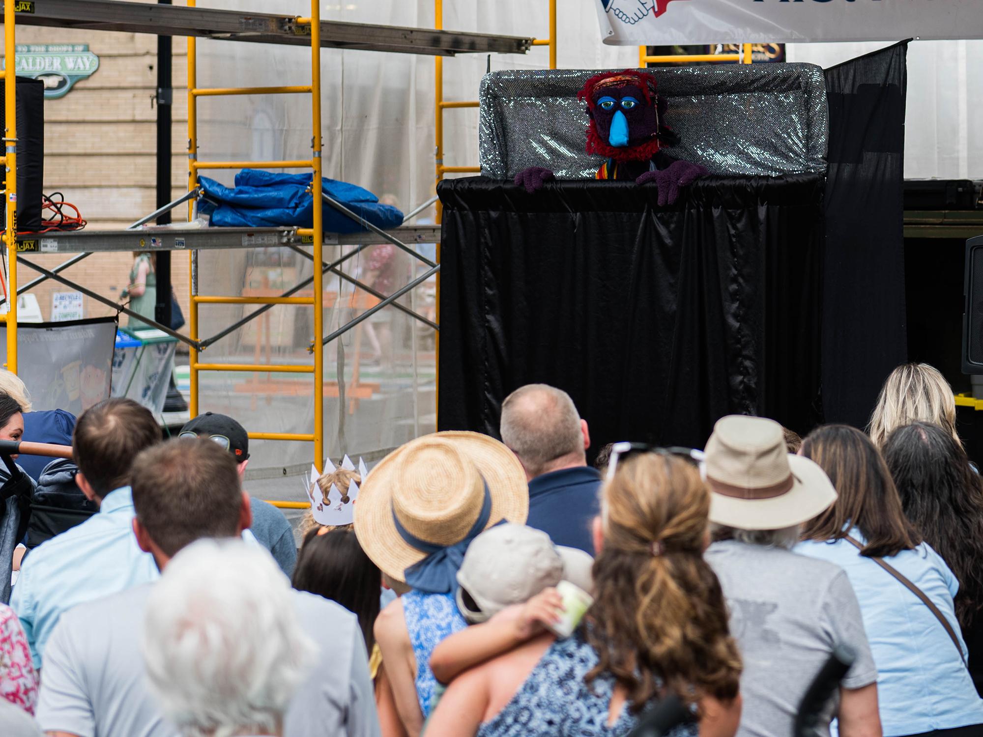A puppet on stage speaks to a gathered audience