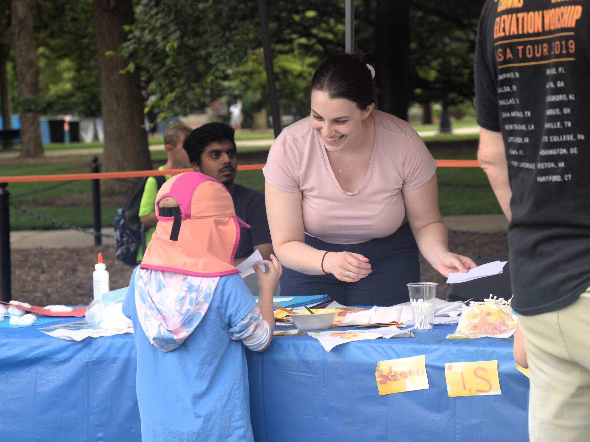 A woman in a pink shirt leans to talk to a kid in front of her table