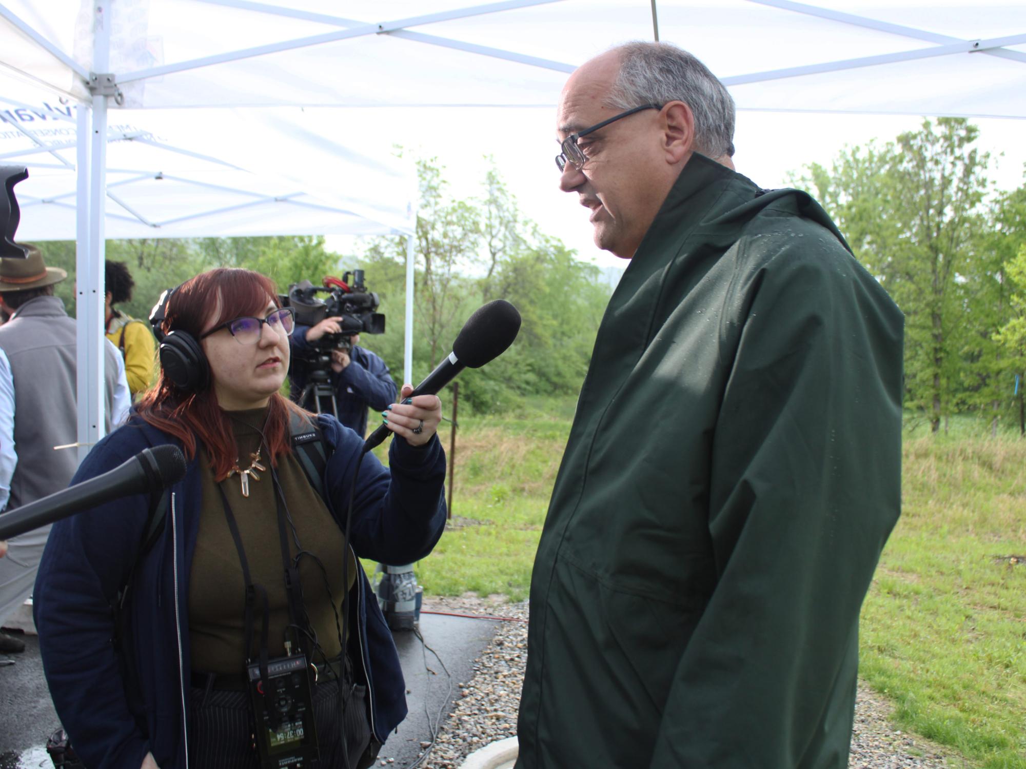 Radio reporter holding a microphone interviewing someone in a field on a cloudy day.