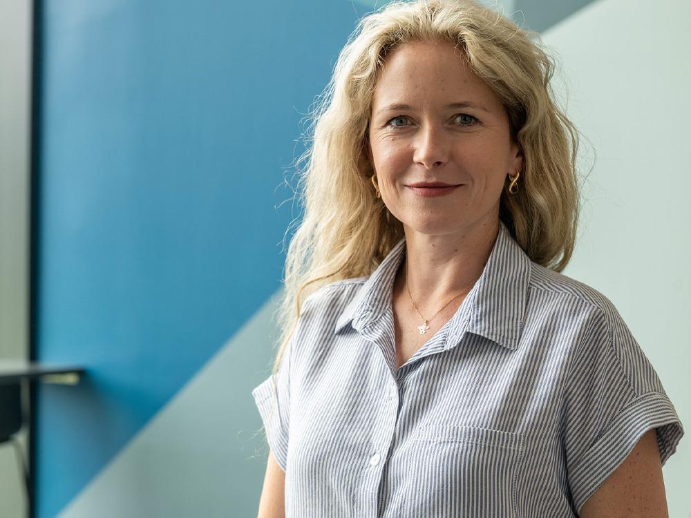 Person looking at the camera, smiling. Inside a building with a blue back wall.