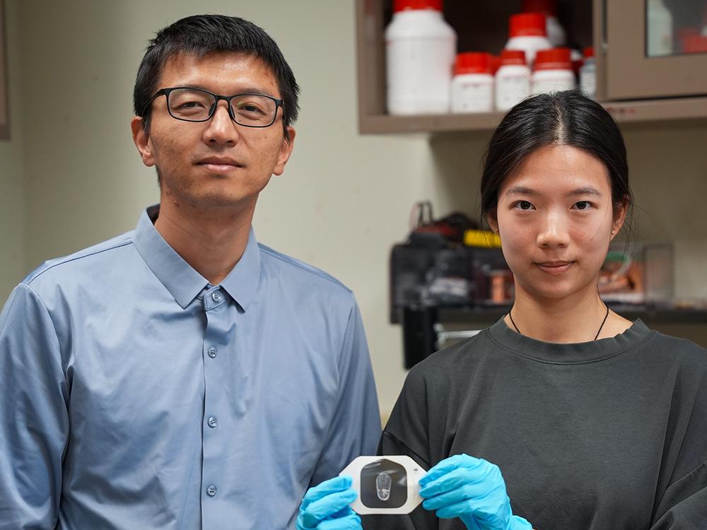 Two researchers smiling at the camera, standing in a lab and holding up their sensor.