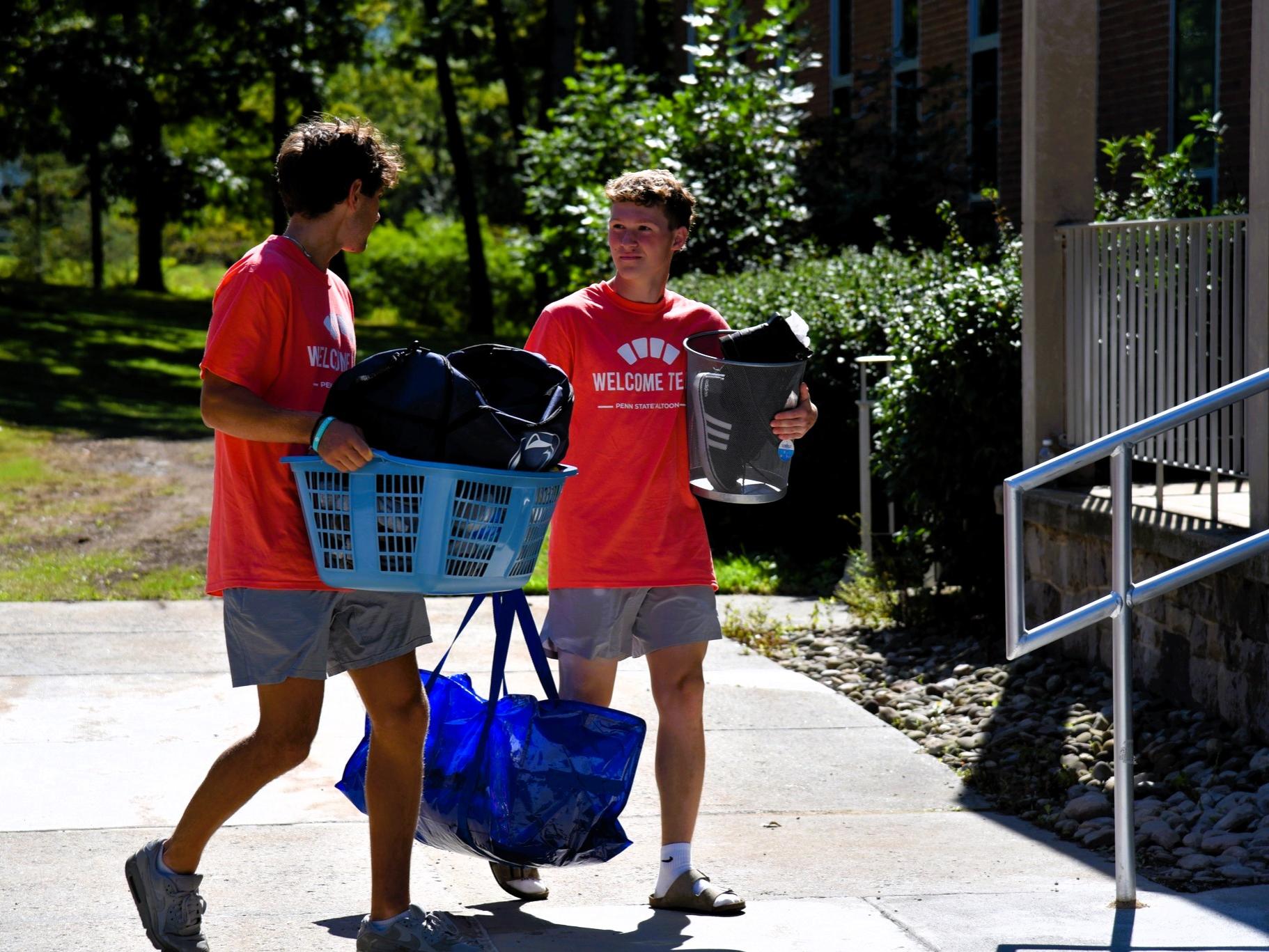 Two students in orange t-shirts carry items into a residence hall