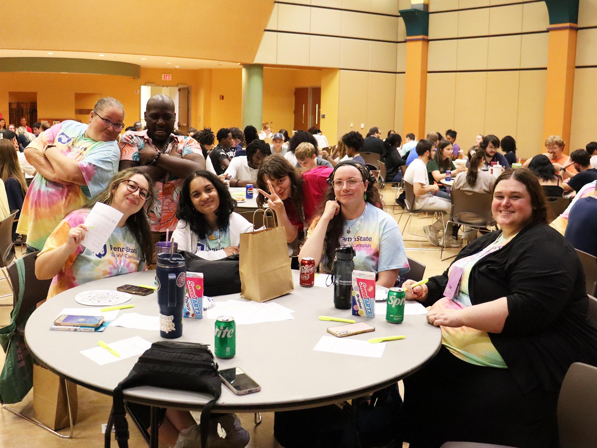 Seven people wearing tie-dye shirts sit around a table in a large room.