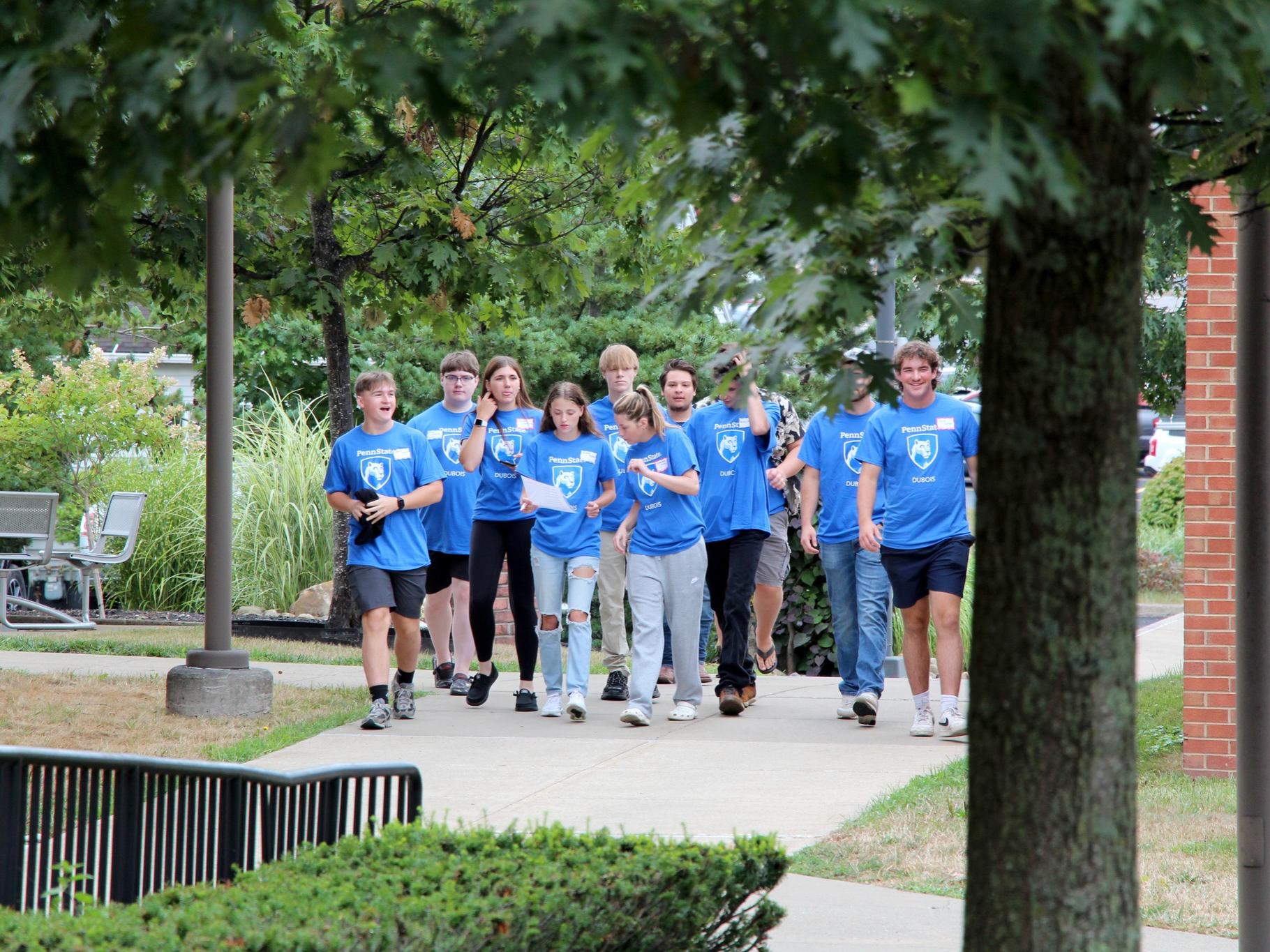 A group of students wearing light blue shirts walk on a college campus