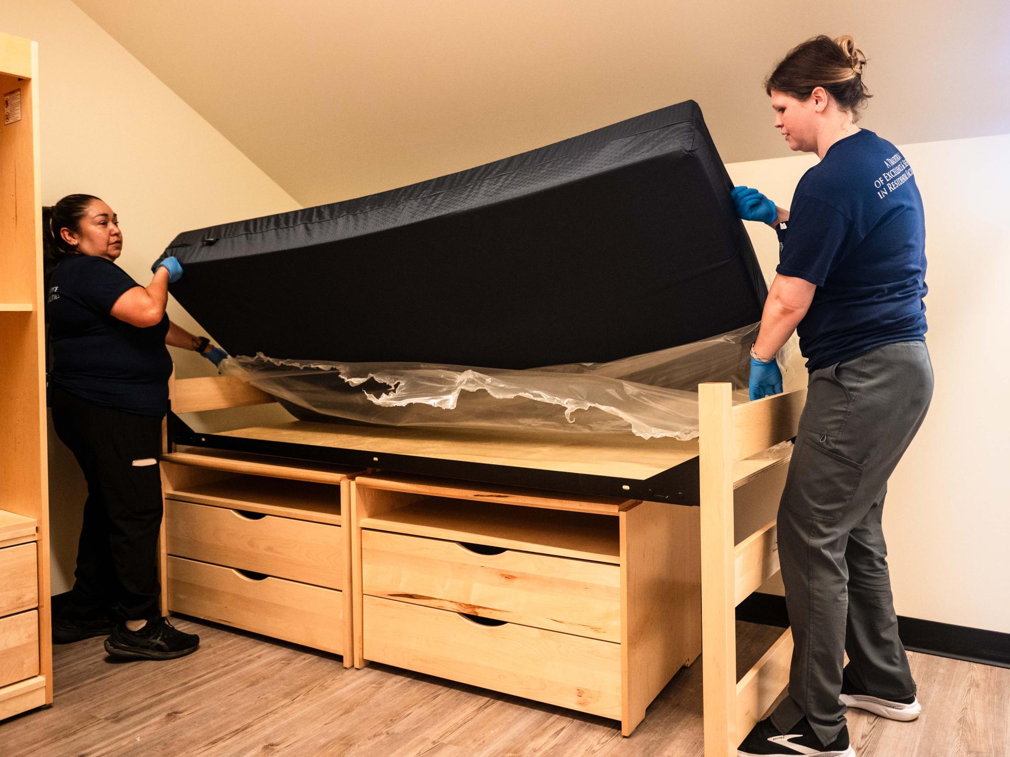 Two staff members place a new mattress on a dorm room bed